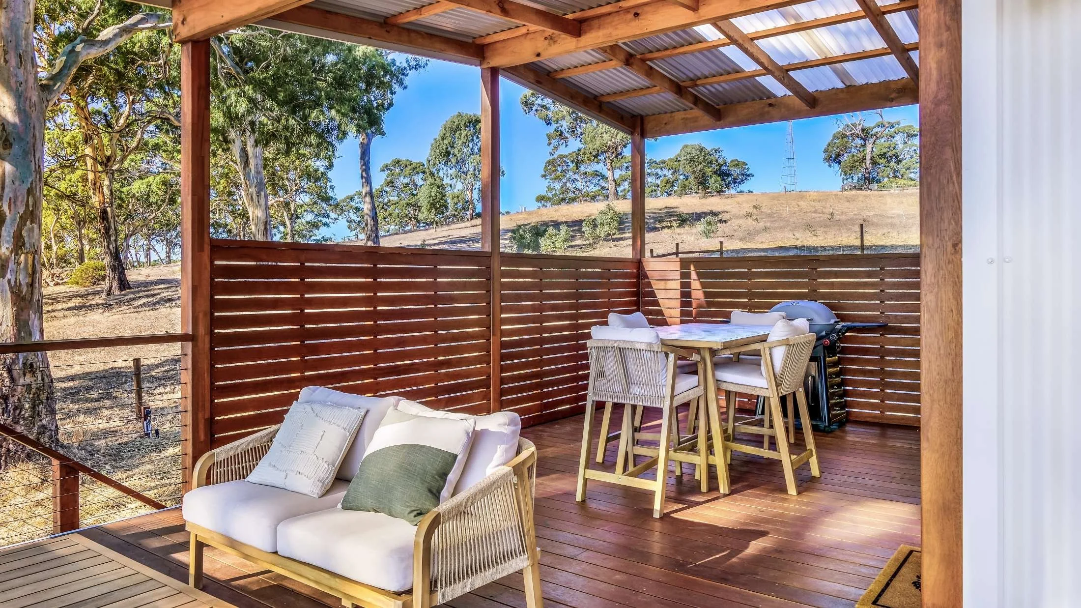 Outdoor patio with a wooden deck, seating area with cushions, and a bar-height table with four chairs, surrounded by a railing, overlooking a hillside with trees and clear blue sky.