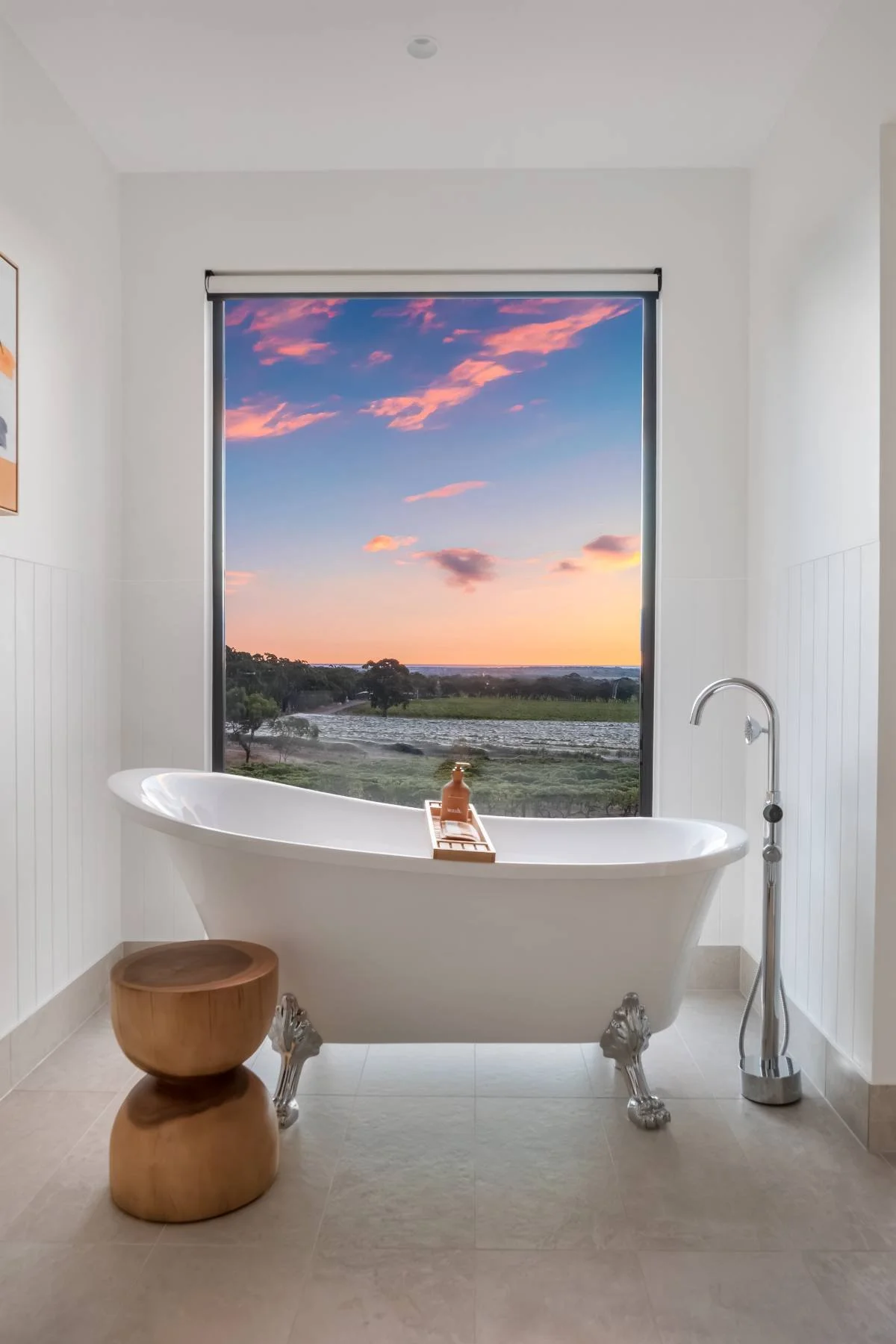 A bathroom with a white clawfoot bathtub, a small wooden stool, and a floor-mounted shower head next to a large window showing a scenic landscape with a sunset sky, trees, and a water body.