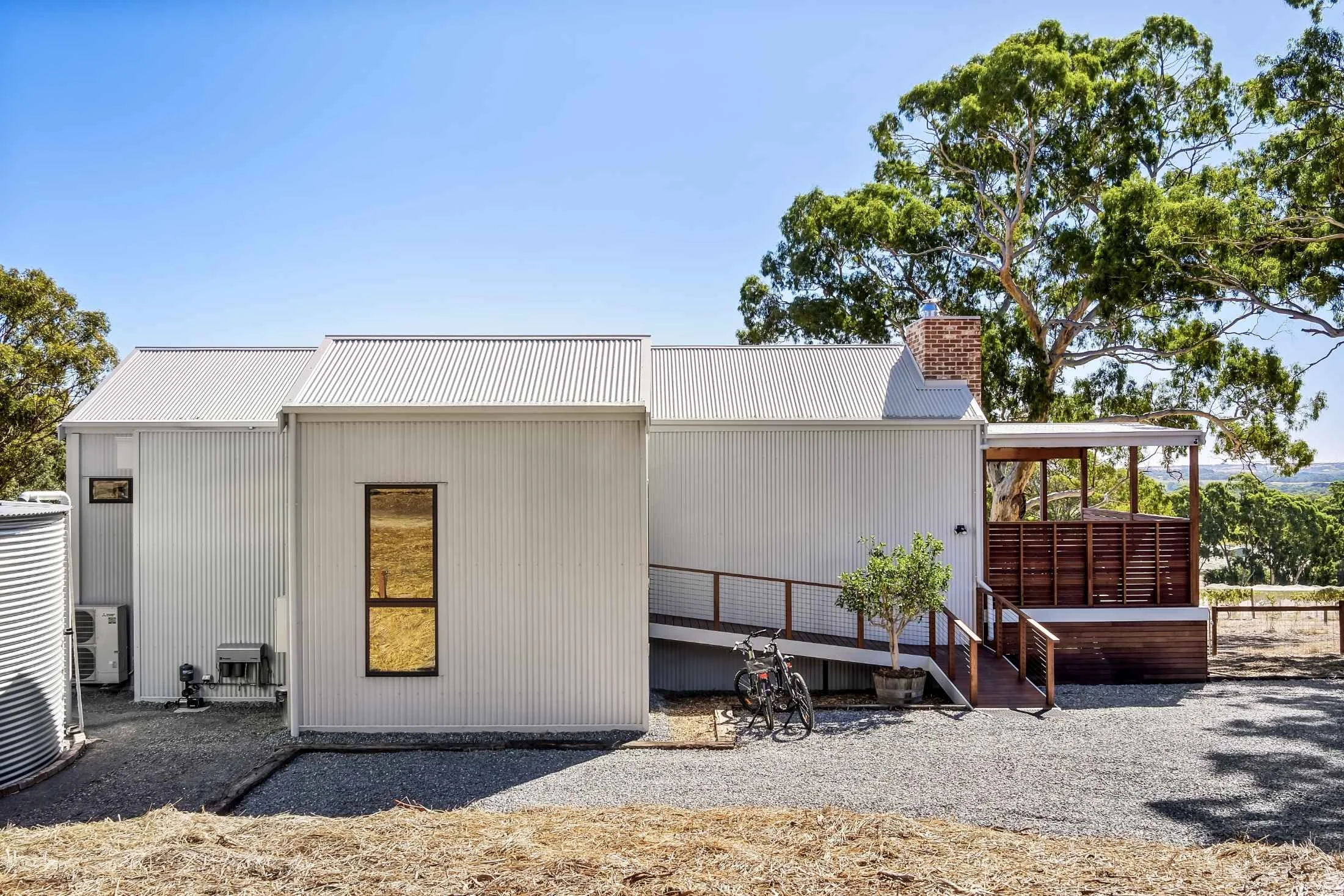 A modern, white, corrugated metal house with a metal roof and a brick chimney. There is a small wooden deck with a ramp, a potted plant, and a bicycle in front. The house is surrounded by trees and gravel ground under a clear blue sky.