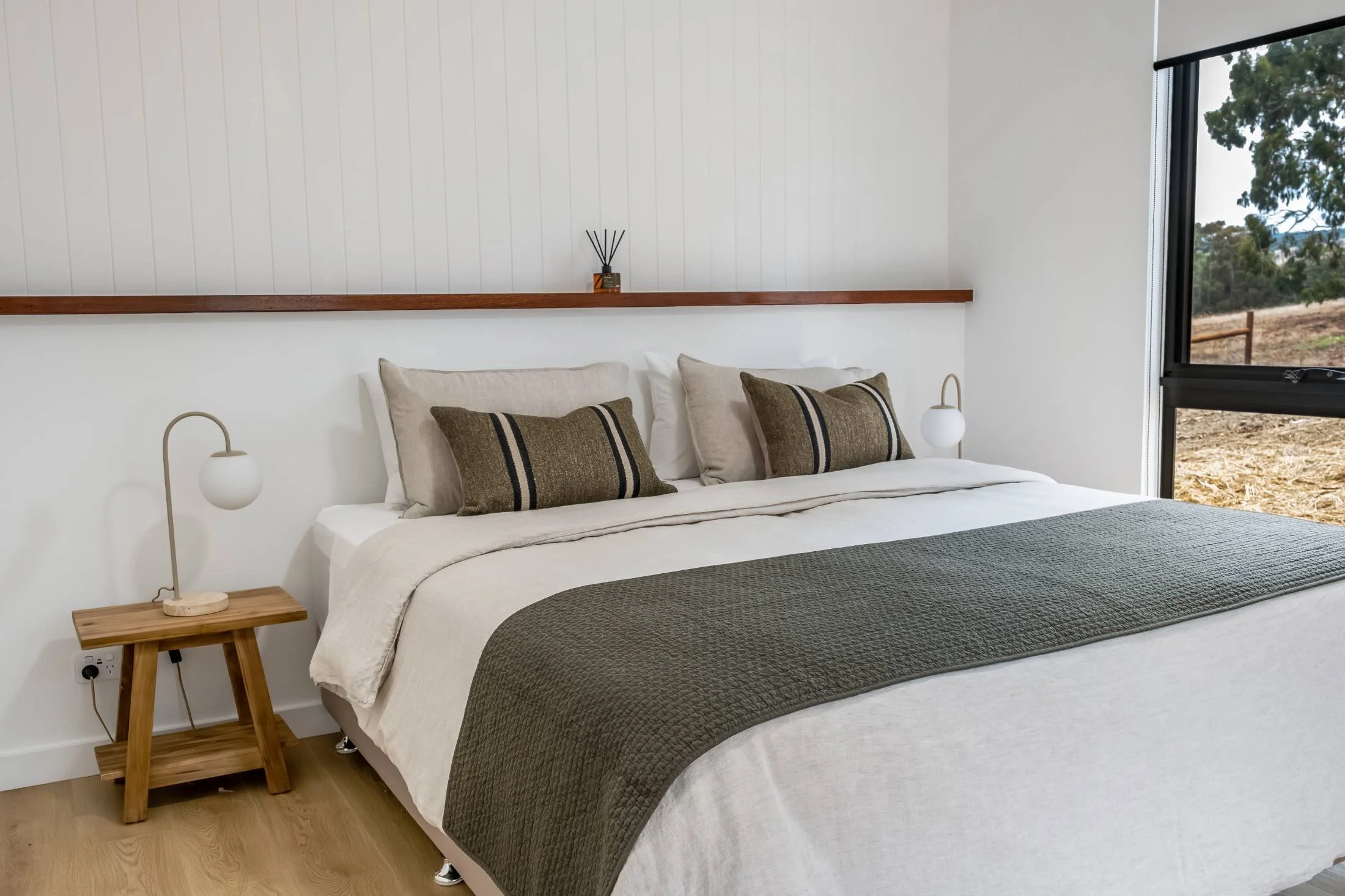 A neatly made bed with beige and brown pillows, a dark green quilt, and white sheets, located in a bright room with a large window showing an outdoor view.