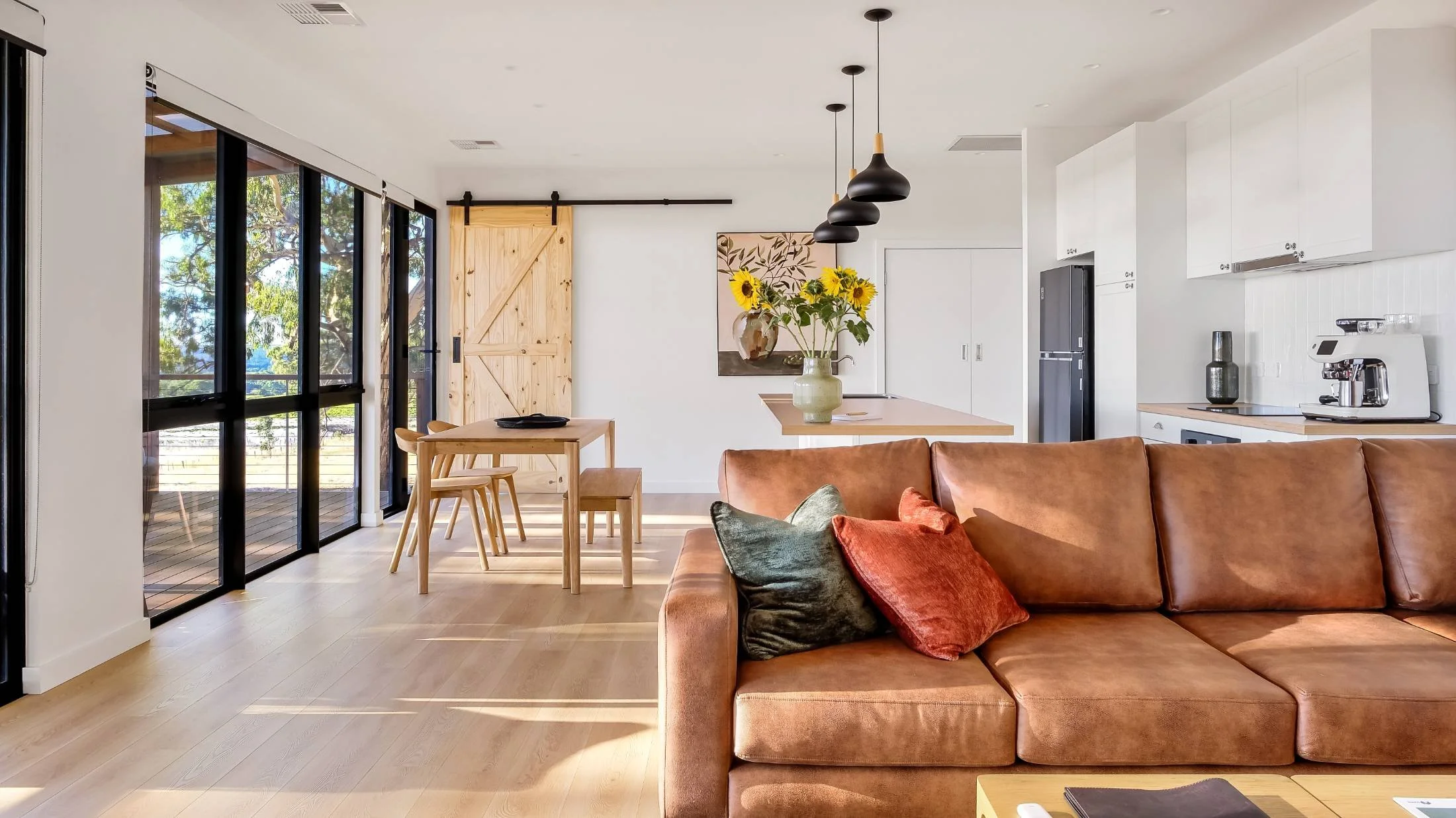 Open-concept living space with a brown sofa, colorful pillows, a wooden dining table with chairs, a sliding glass door leading to an outdoor deck, a kitchen area with white cabinets, a black refrigerator, and pendant lights hanging above a vase with 