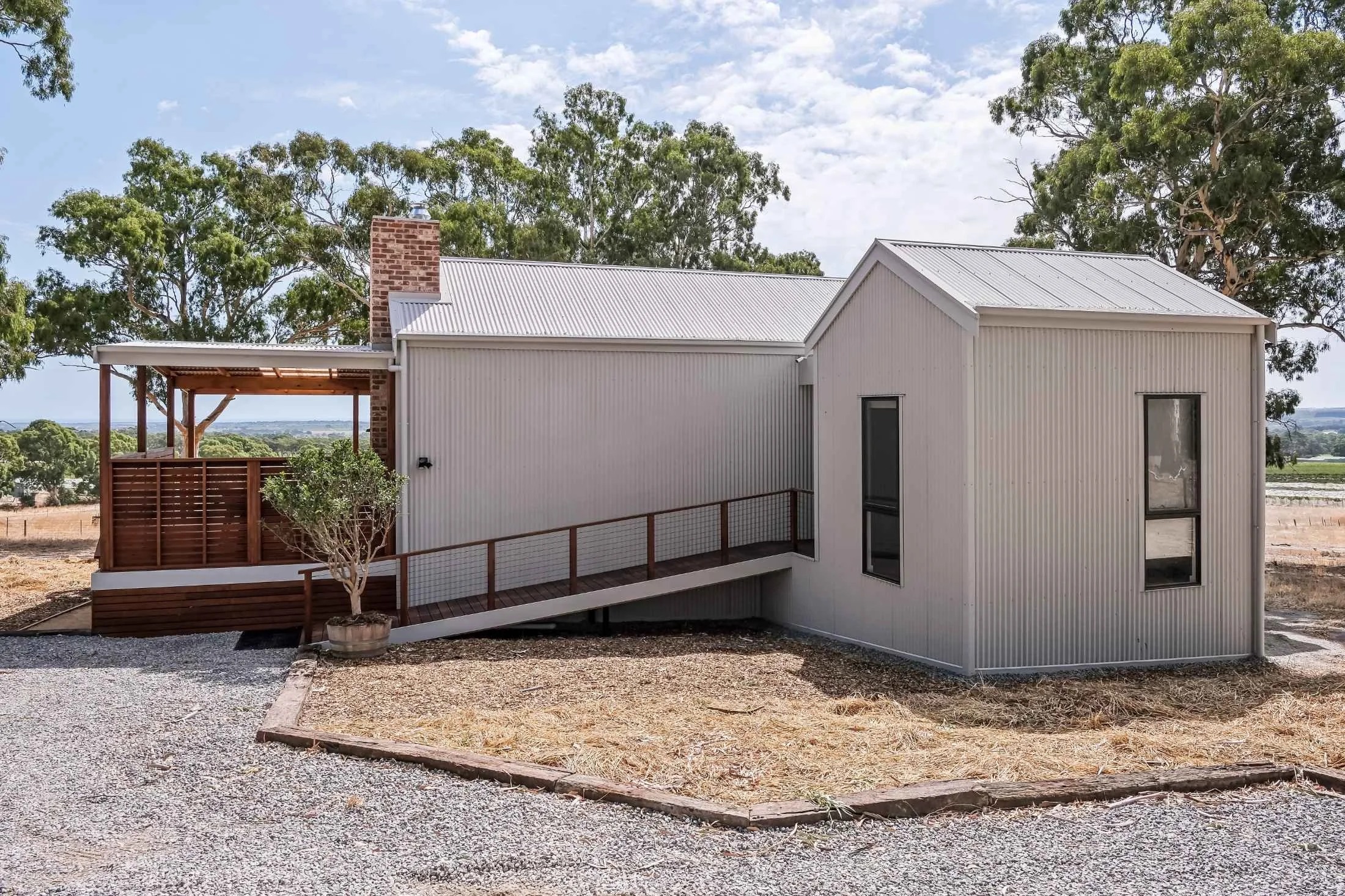 Modern house with metal siding, two large windows, a brick chimney, and a small wooden deck with a railing. The house is situated on a gravel yard with trees in the background.