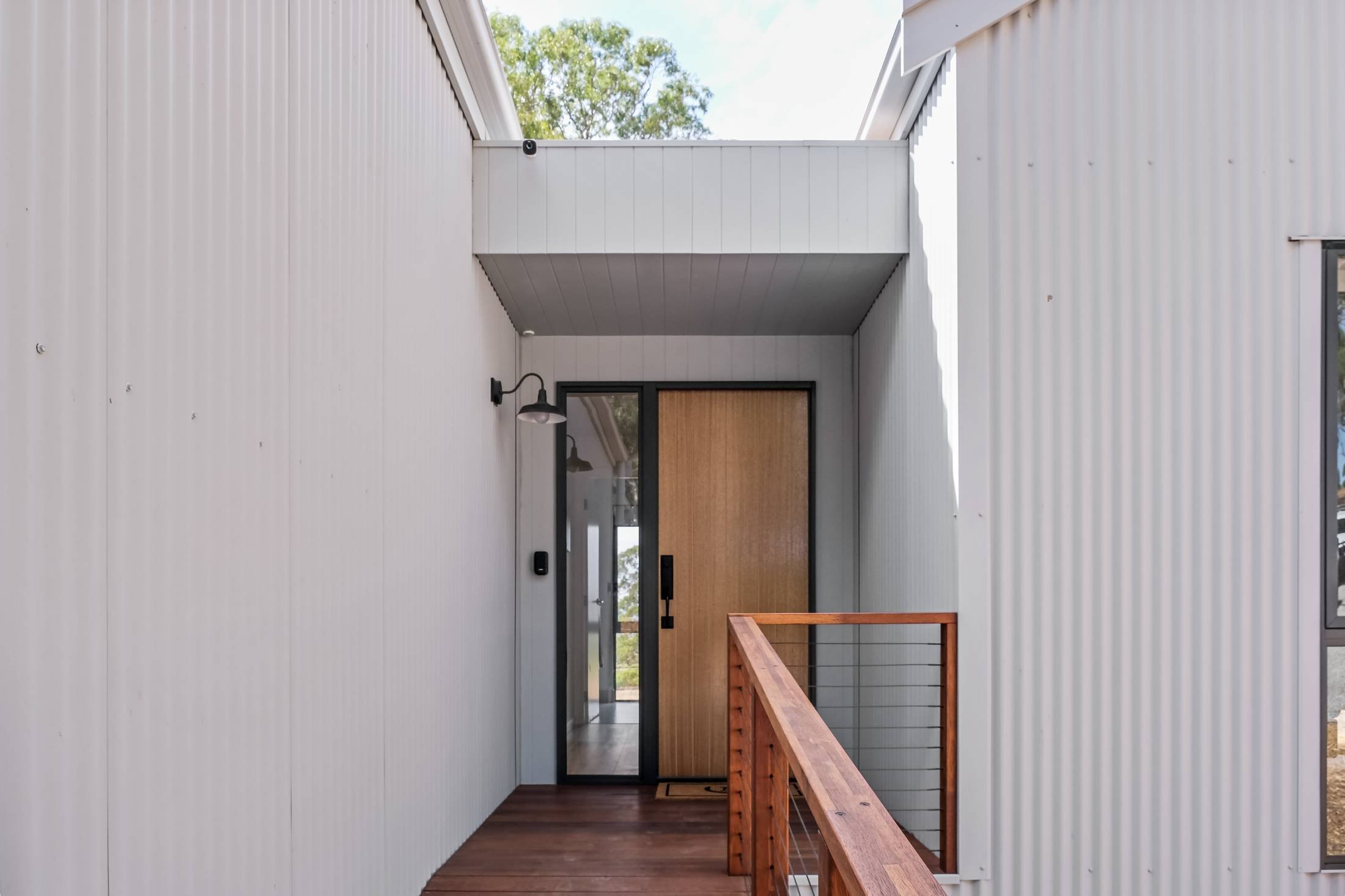 Front porch area of a modern house with white vertical siding, a wooden door, a black-framed glass panel, a black wall-mounted light fixture, a wooden railing, and a partial second floor deck overhead.
