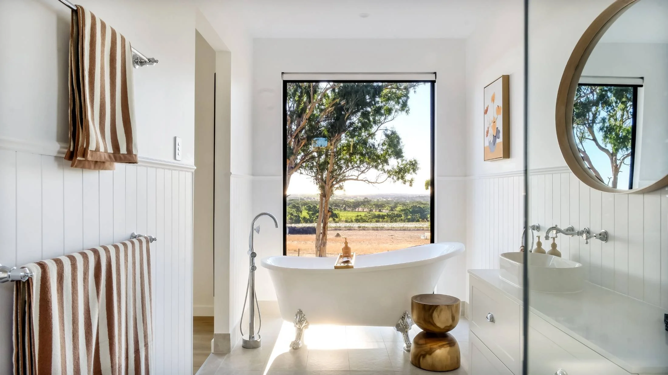 Bright bathroom with a freestanding clawfoot bathtub positioned in front of a large window showing an outdoor landscape with trees and open sky. A wooden stool is next to the bathtub. The bathroom has white walls with beadboard paneling, a white vani