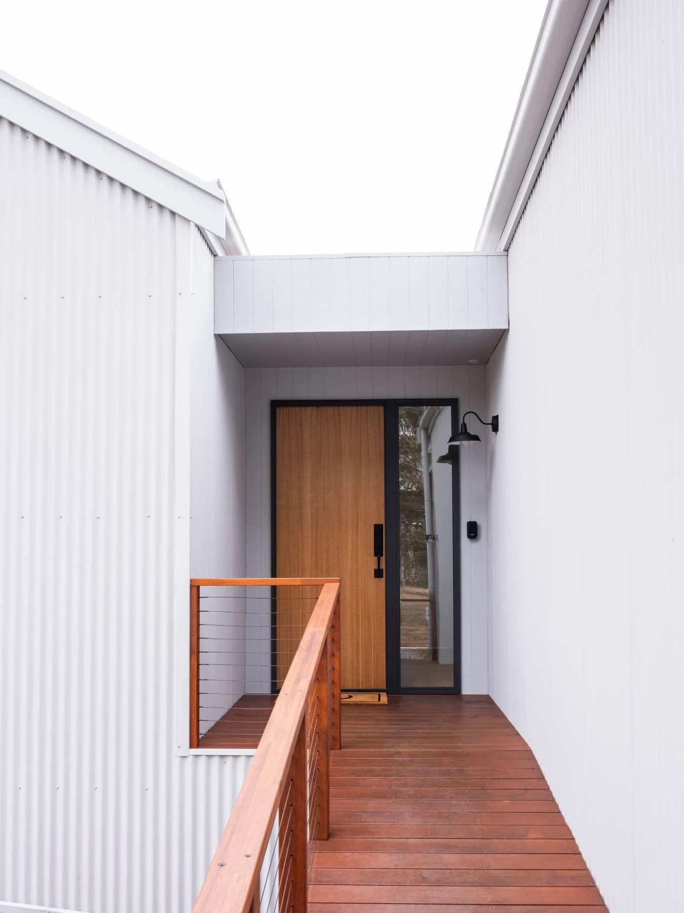 Exterior front porch of a modern home with white corrugated siding, a wooden front door, and a wooden railing, with a black outdoor light fixture.