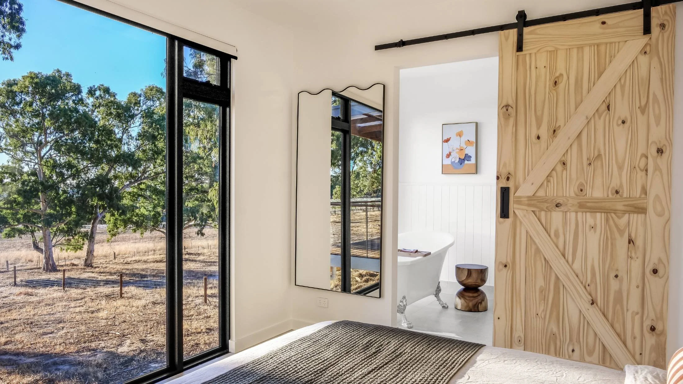 View from inside a modern bedroom with a large sliding glass door opening to a natural outdoor scene with trees and dry grass. Part of a bed with a textured blanket is visible, along with a tall mirror and a wooden sliding barn door leading to a bath