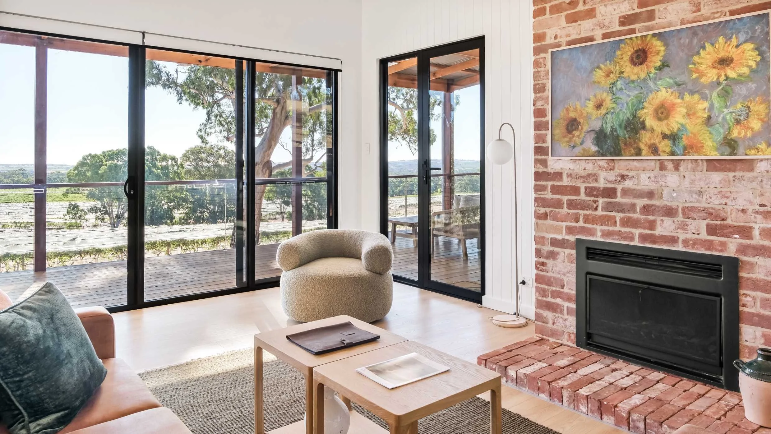 Living room with a brick fireplace, sunflowers painting, beige armchair, sliding glass doors, and outdoor view of trees and sky.