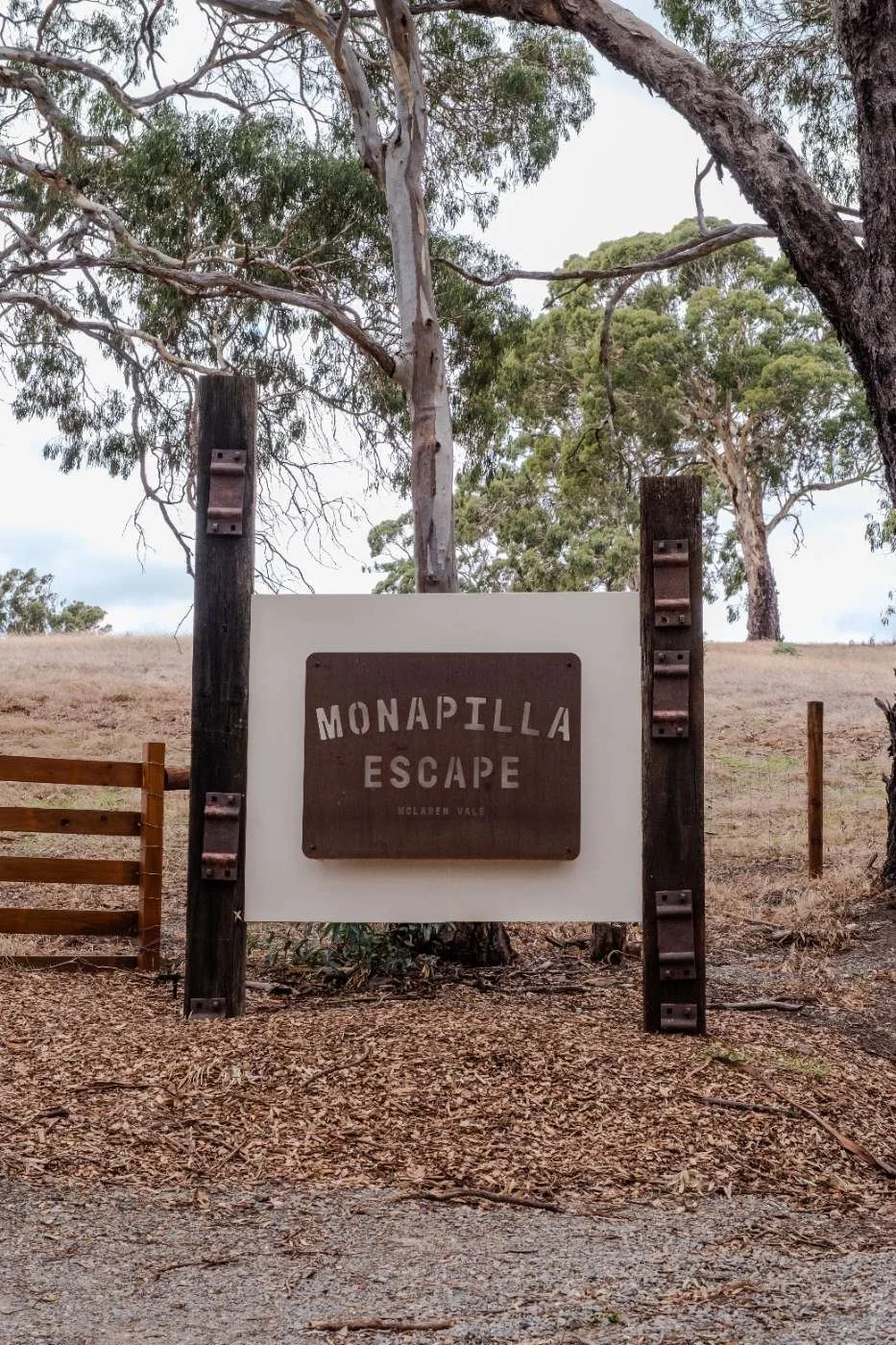 A sign reading 'Monapilla Escape, Molaren Vale' is mounted on a wooden frame in a wooded outdoor area with trees and a dry grassy field in the background.