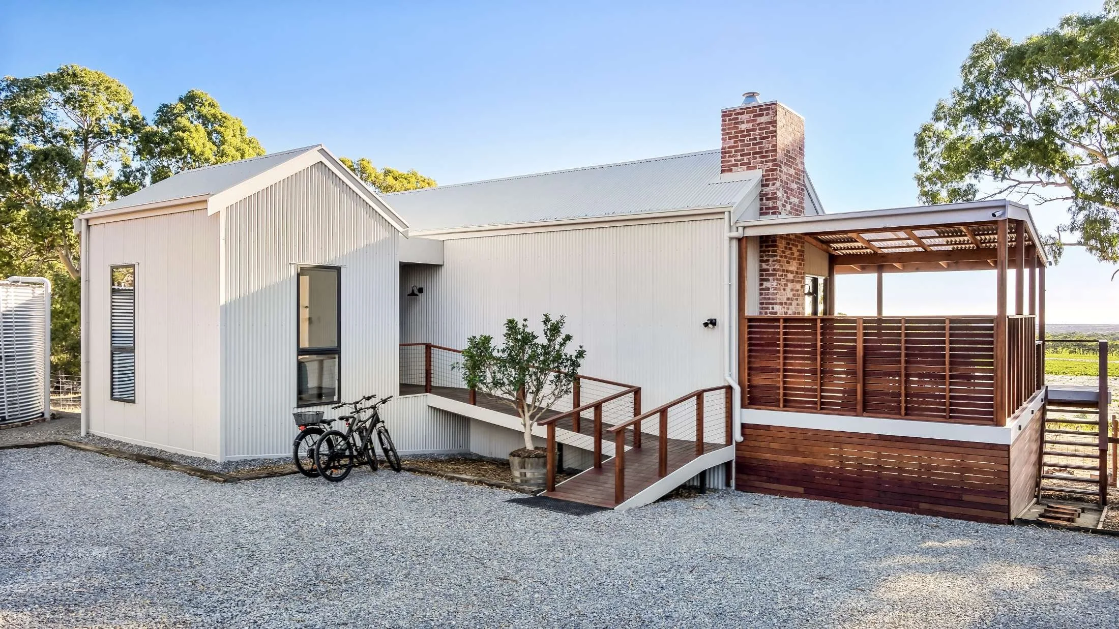 Modern house with white metal siding, a wooden deck with railings, a brick chimney, and two bicycles parked outside. Gravel pathway in front, trees in background, clear blue sky.