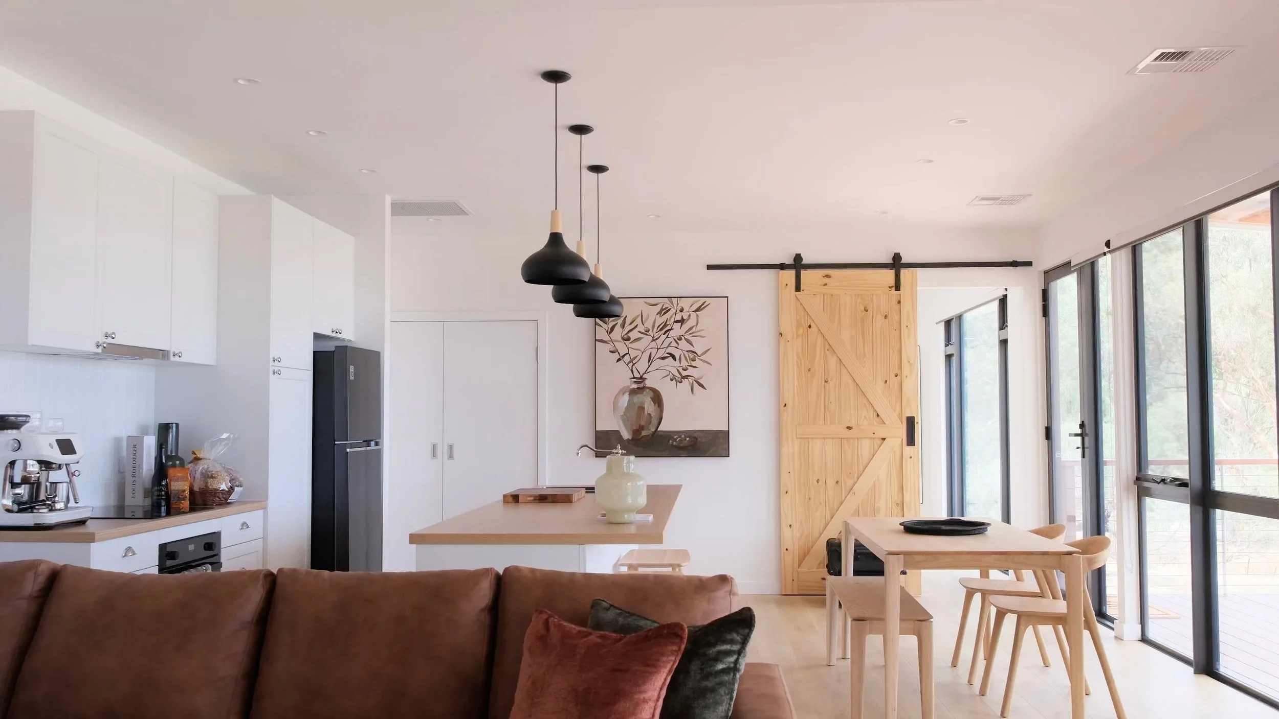Open-concept kitchen and dining area with white cabinets, a wooden sliding barn door, a wooden dining table with chairs, a sofa with cushions, and large windows bringing in natural light.