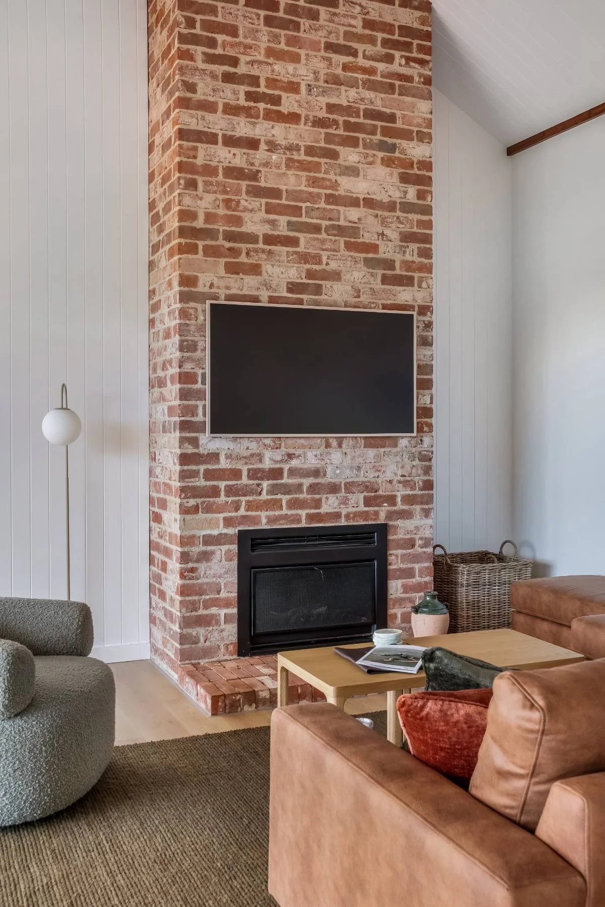 Living room with a brick fireplace, flat-screen TV mounted above, beige couch with a red pillow, a wooden coffee table with magazines and a ceramic vase, a wicker basket, a gray textured armchair, a tall floor lamp, and a neutral-toned rug.