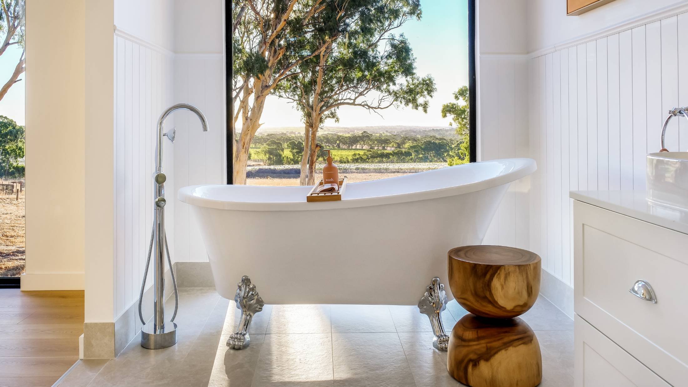 White clawfoot bathtub with silver lion head feet, placed in front of large window showing trees and a landscape, with a wooden side table beside it.