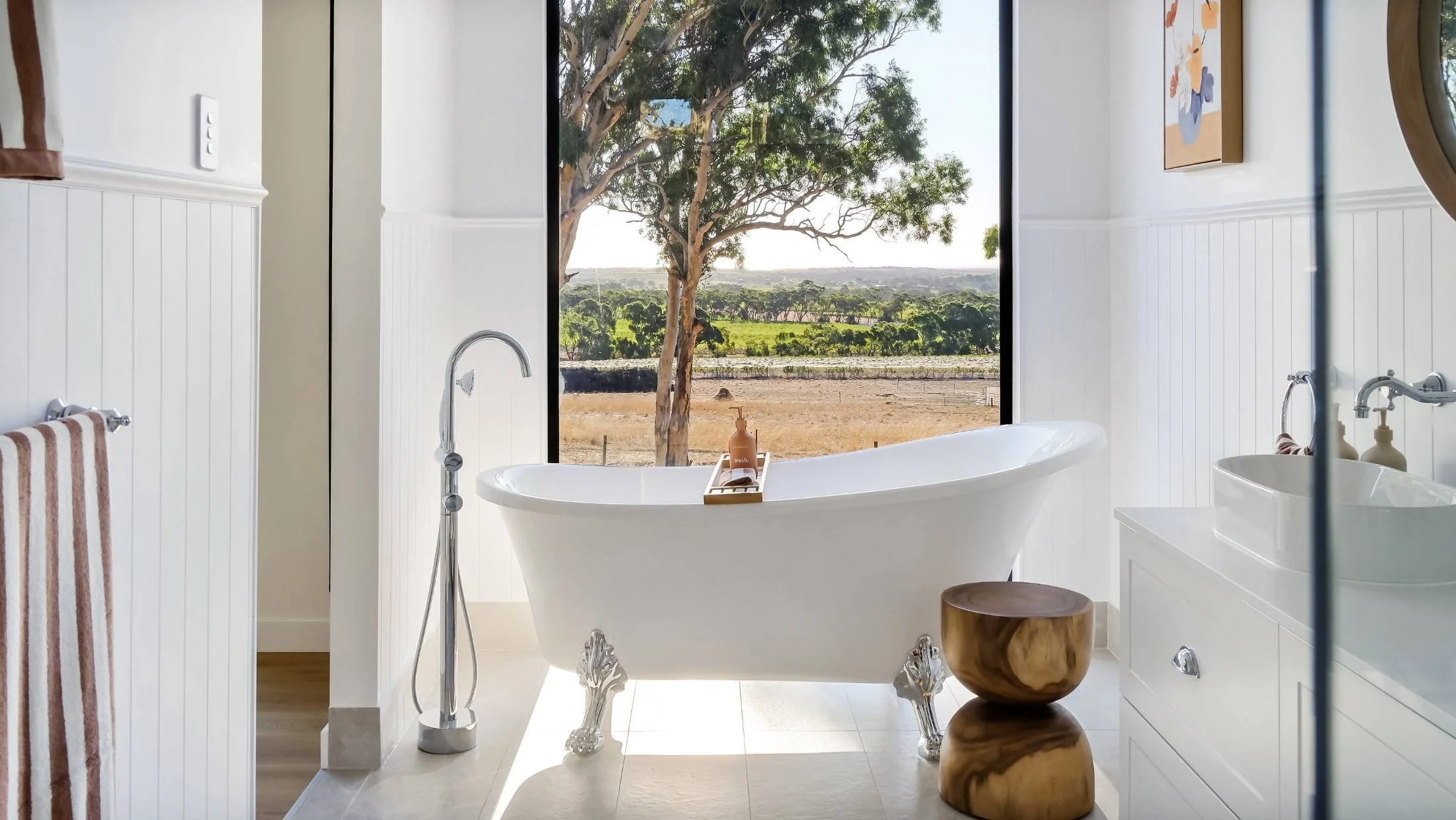 Bathroom with a white clawfoot bathtub in front of large window showing outdoor scenery of trees and fields, with a small wooden stool beside the tub and a white vanity with a vessel sink on the right.
