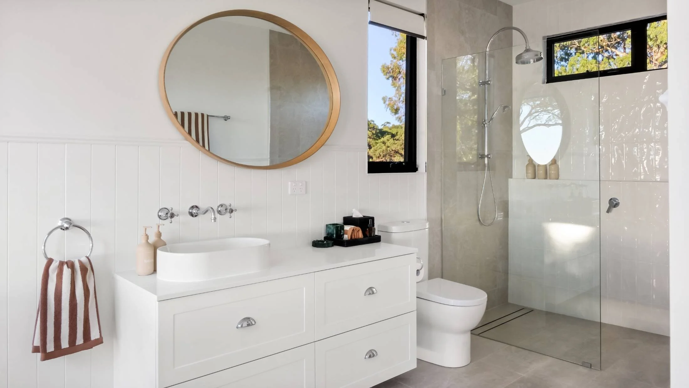 Modern bathroom with white vanity, round mirror, shower with glass door, and window showing outdoor scenery.