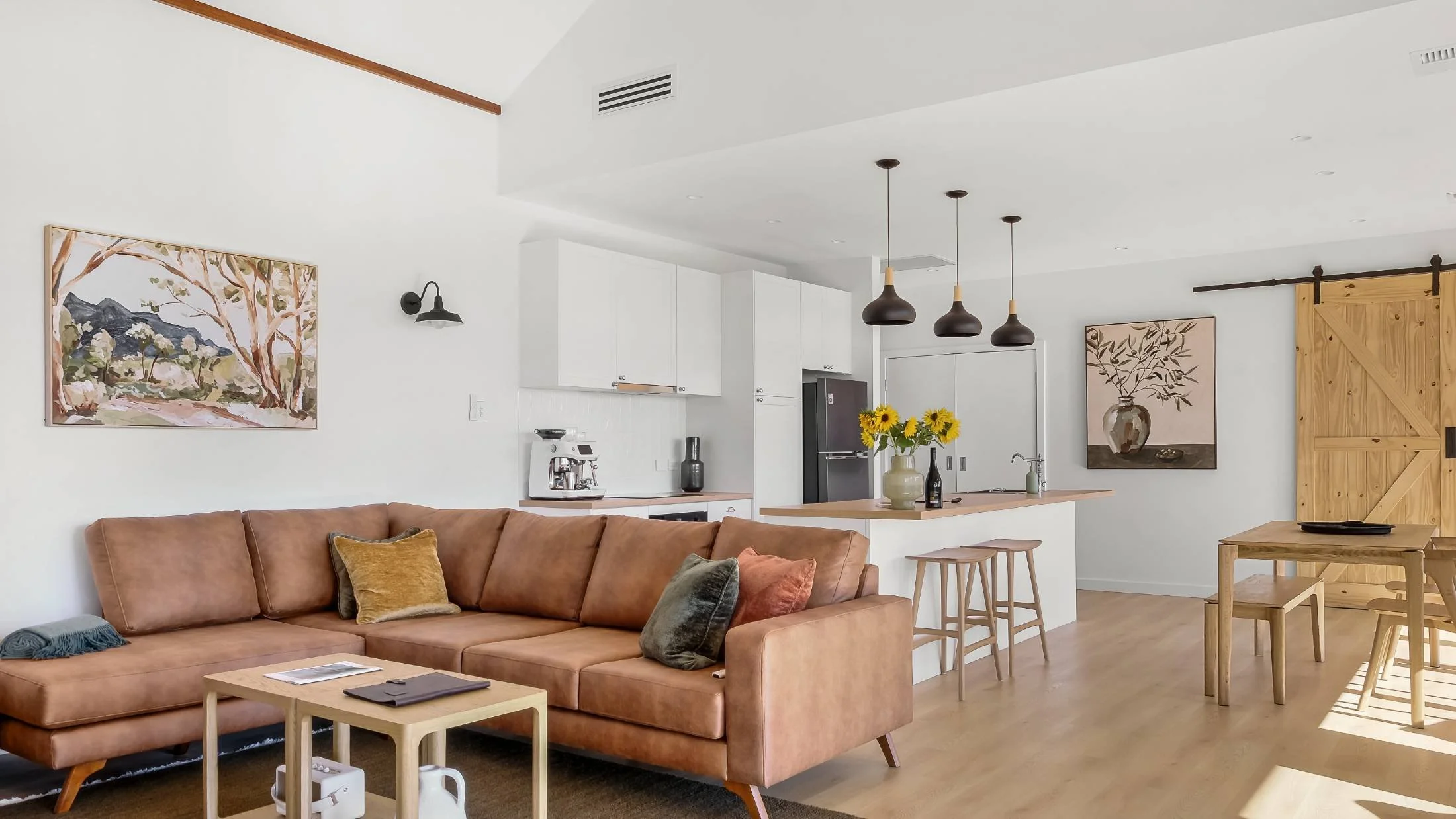 Open-concept living room and kitchen with a tan sectional, wood coffee table, three black pendant lights, white cabinetry, black refrigerator, sliding barn door, wall art, sunflower decoration, and wooden dining table with chairs.