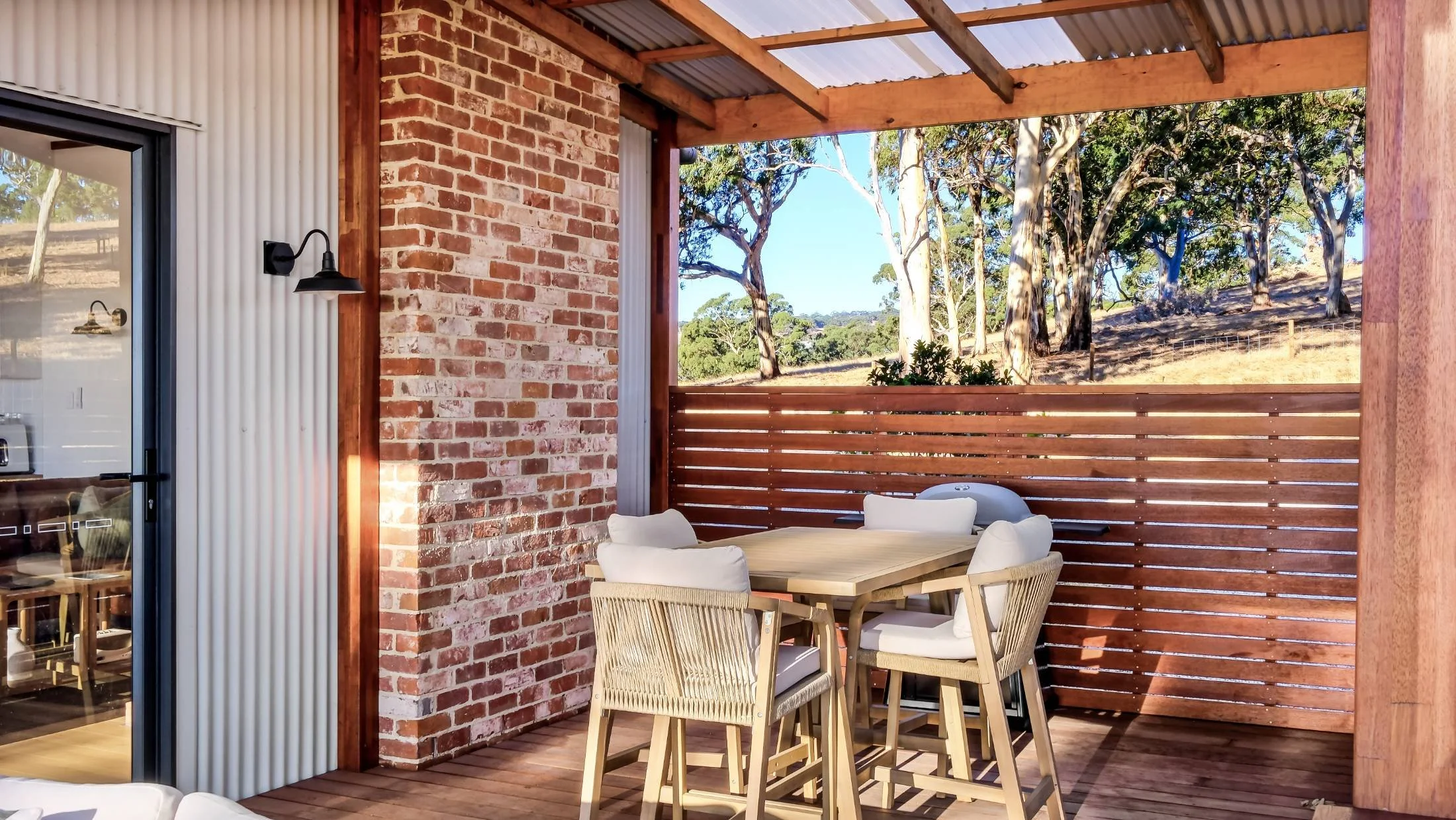 Covered outdoor patio with a wooden dining table and chairs, brick and wood wall, and a view of trees and a hillside.