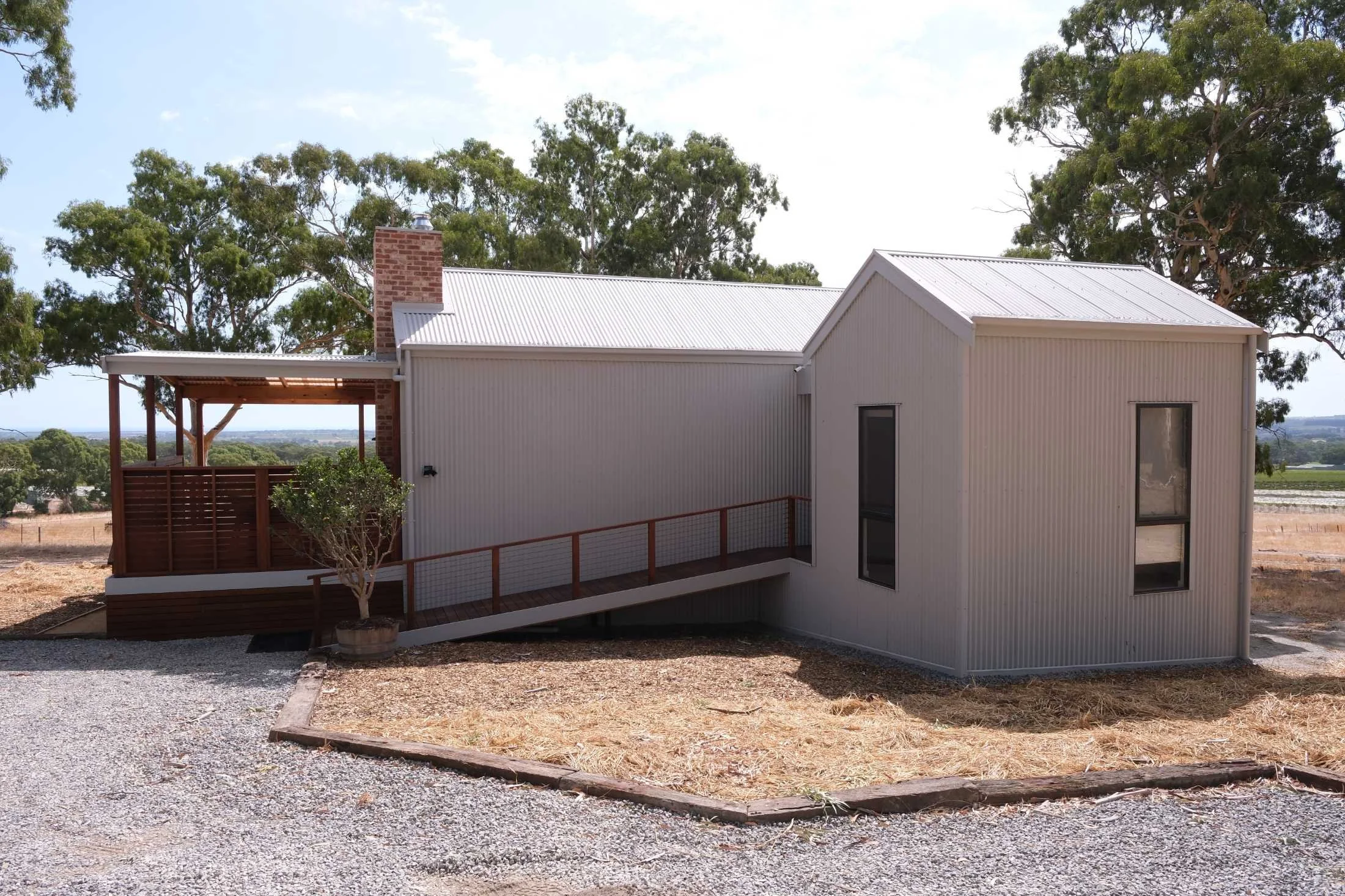 Modern house with metal siding, a wrap-around balcony, and large windows, surrounded by trees and a gravel yard.