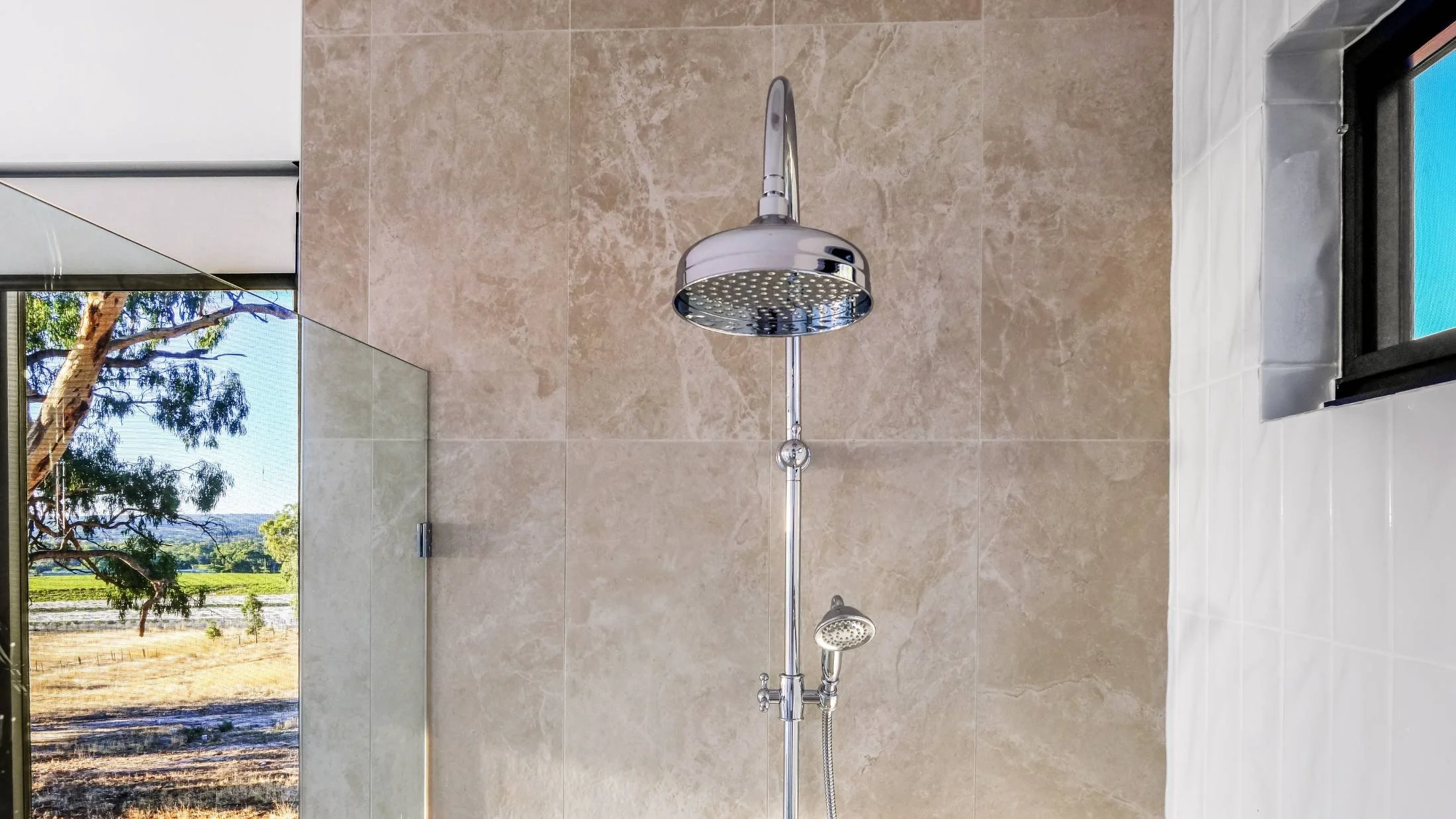 Showerhead and handheld shower fixture on beige tiled wall with a window showing outdoor trees and landscape.