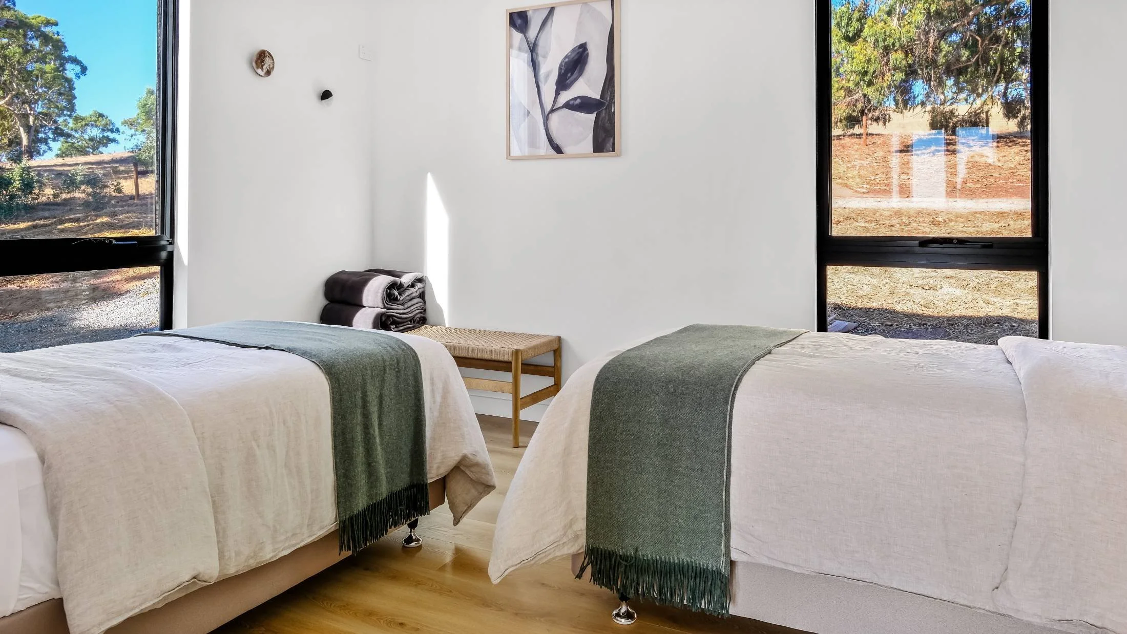 A bedroom with two single beds each with a green blanket, a small wooden bench, folded towels, and large windows showing an outdoor natural scene.