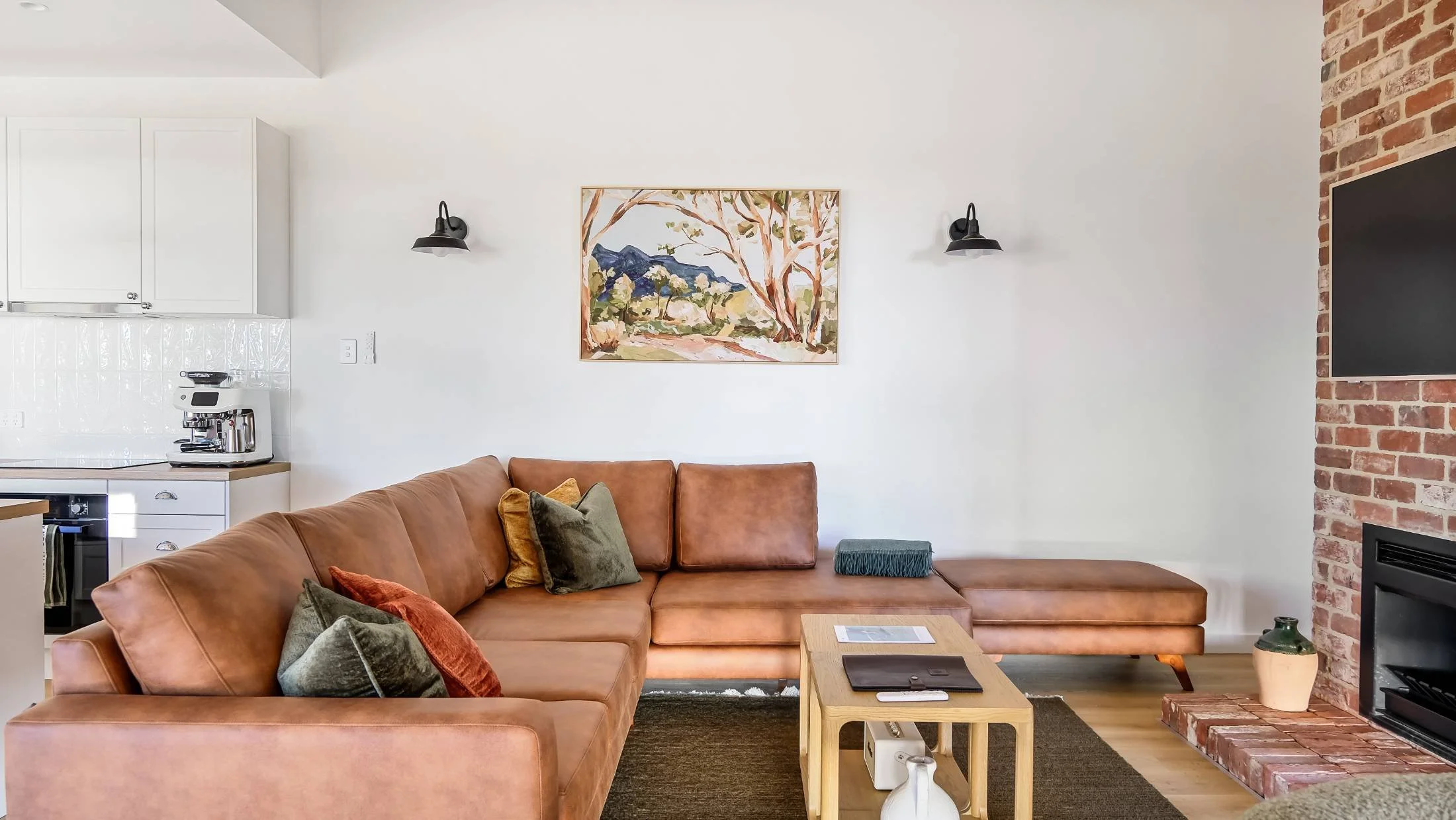 Living room with a brown sectional sofa, a small wooden coffee table, a mounted TV, a brick fireplace, wall-mounted lights, and a painting of mountains and trees.
