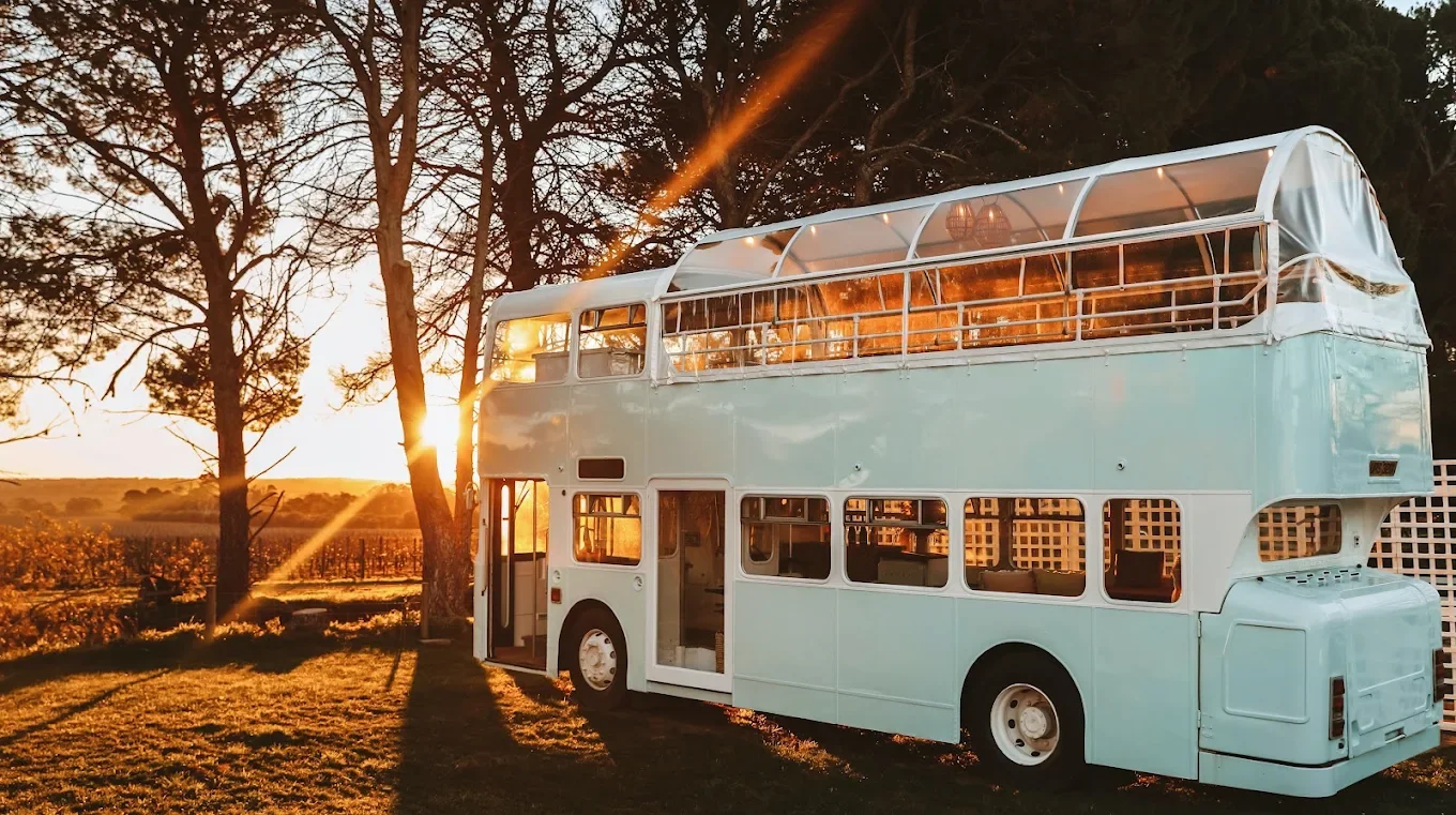 A vintage double-decker bus parked outdoors at sunset, surrounded by trees and vineyards.