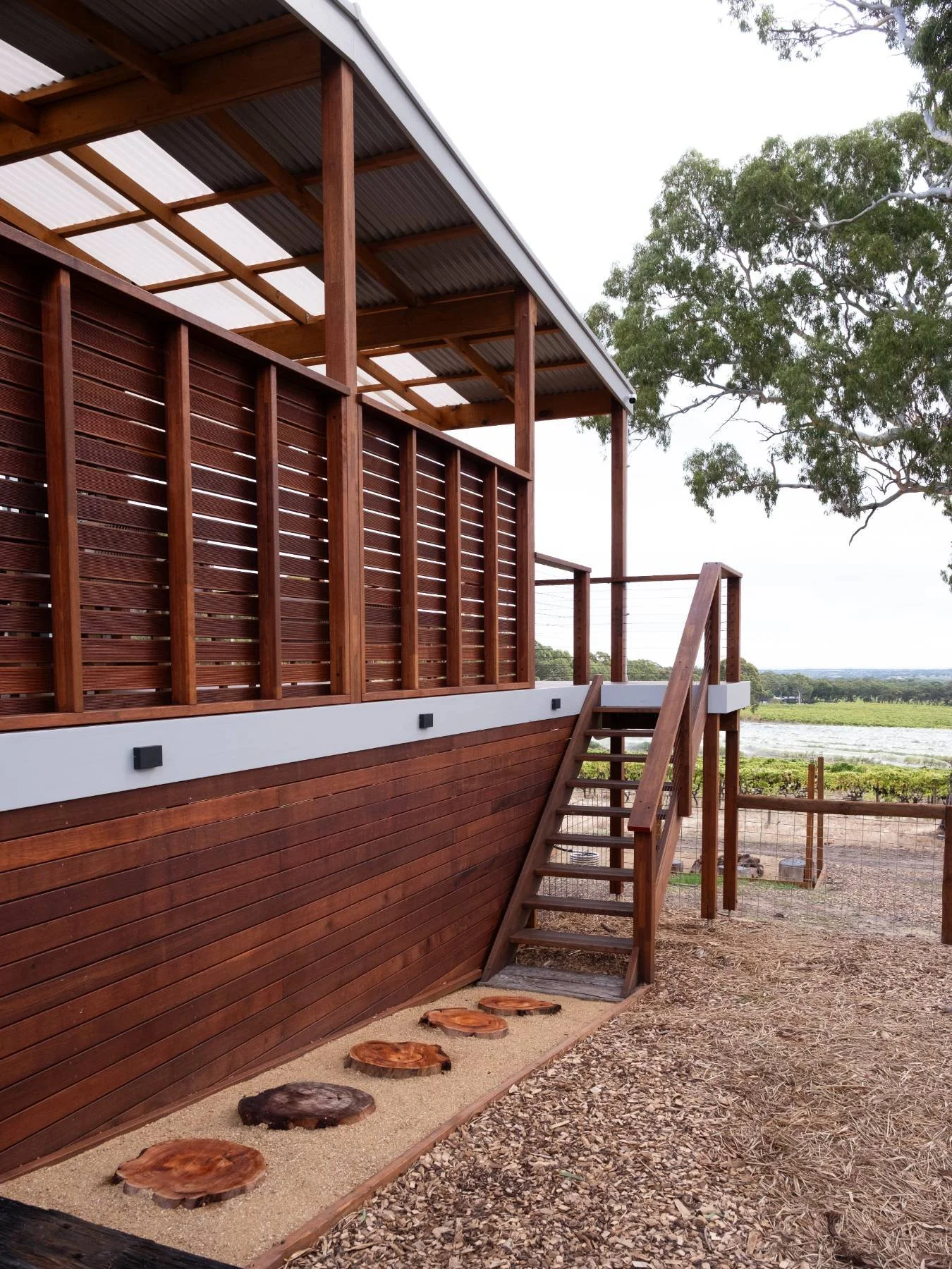 Wooden deck with stairs and a fenced railing, overlooking a rural landscape with fields and trees.