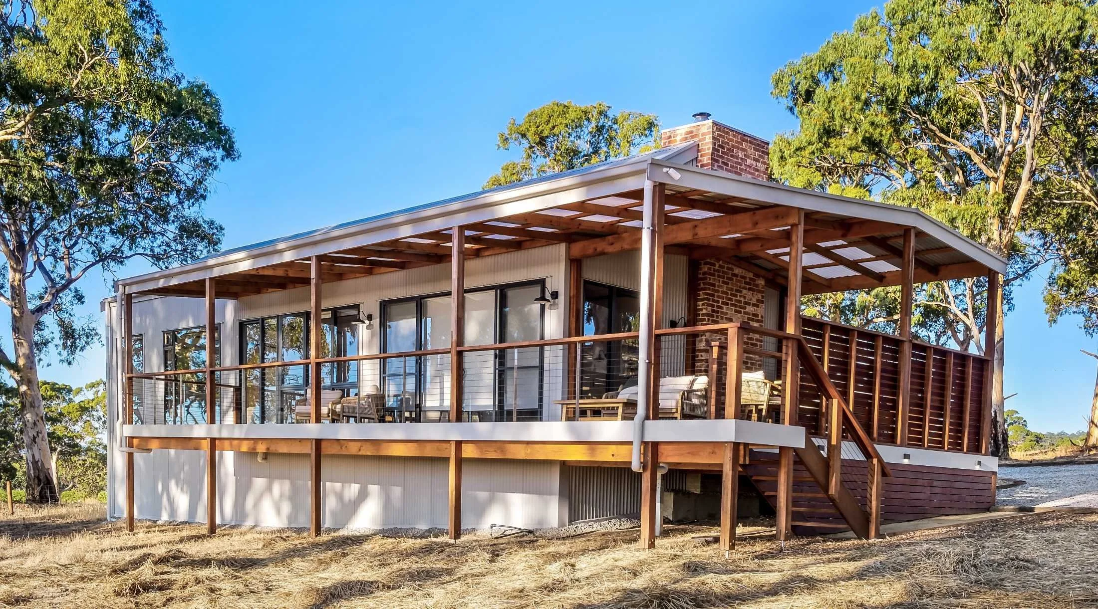 A modern house with a wooden balcony and large glass sliding doors, surrounded by trees under a clear blue sky.