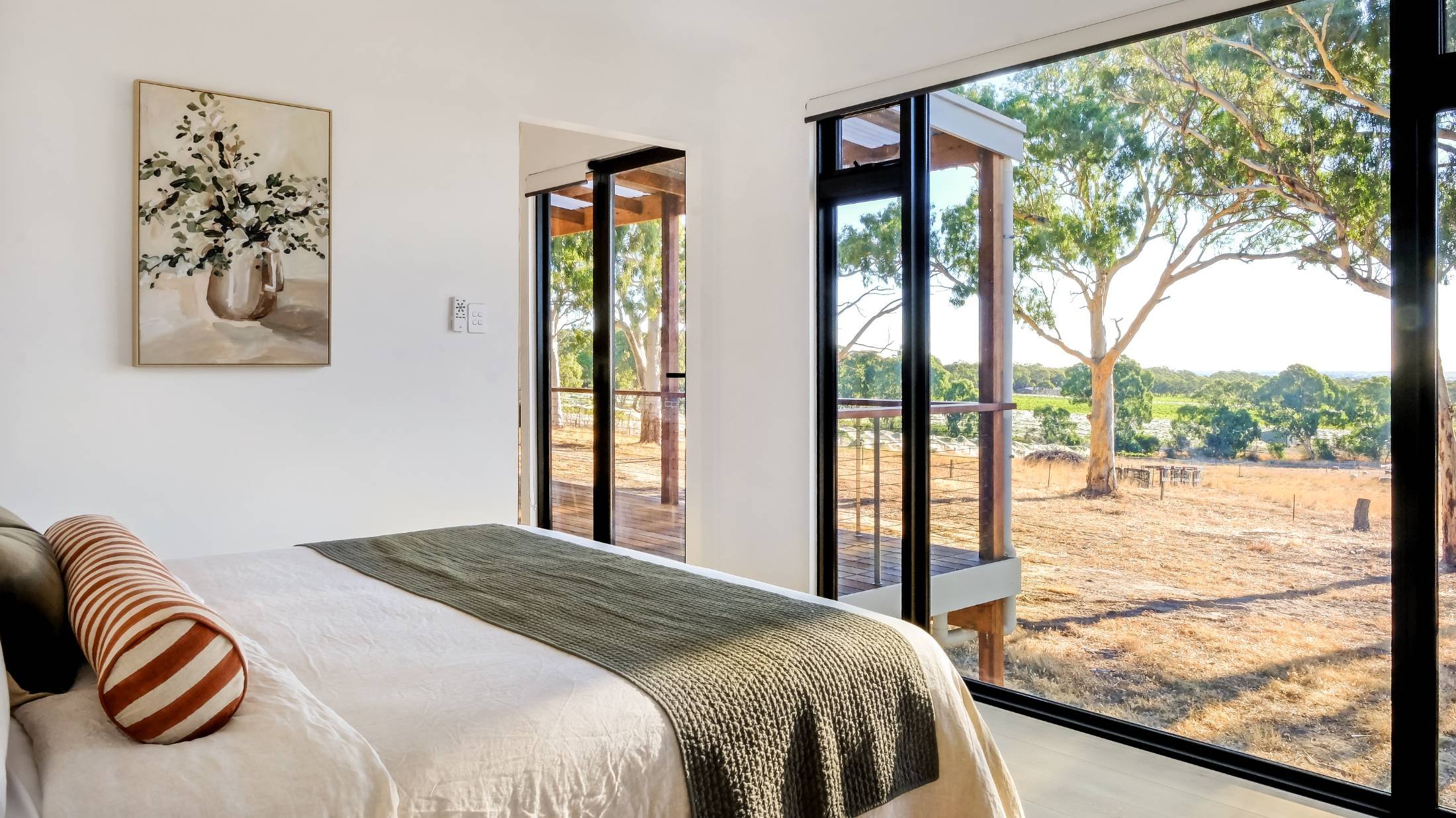 Bedroom with a large window showing an outdoor view of trees and a field, decorated with a bed and artwork on the wall.