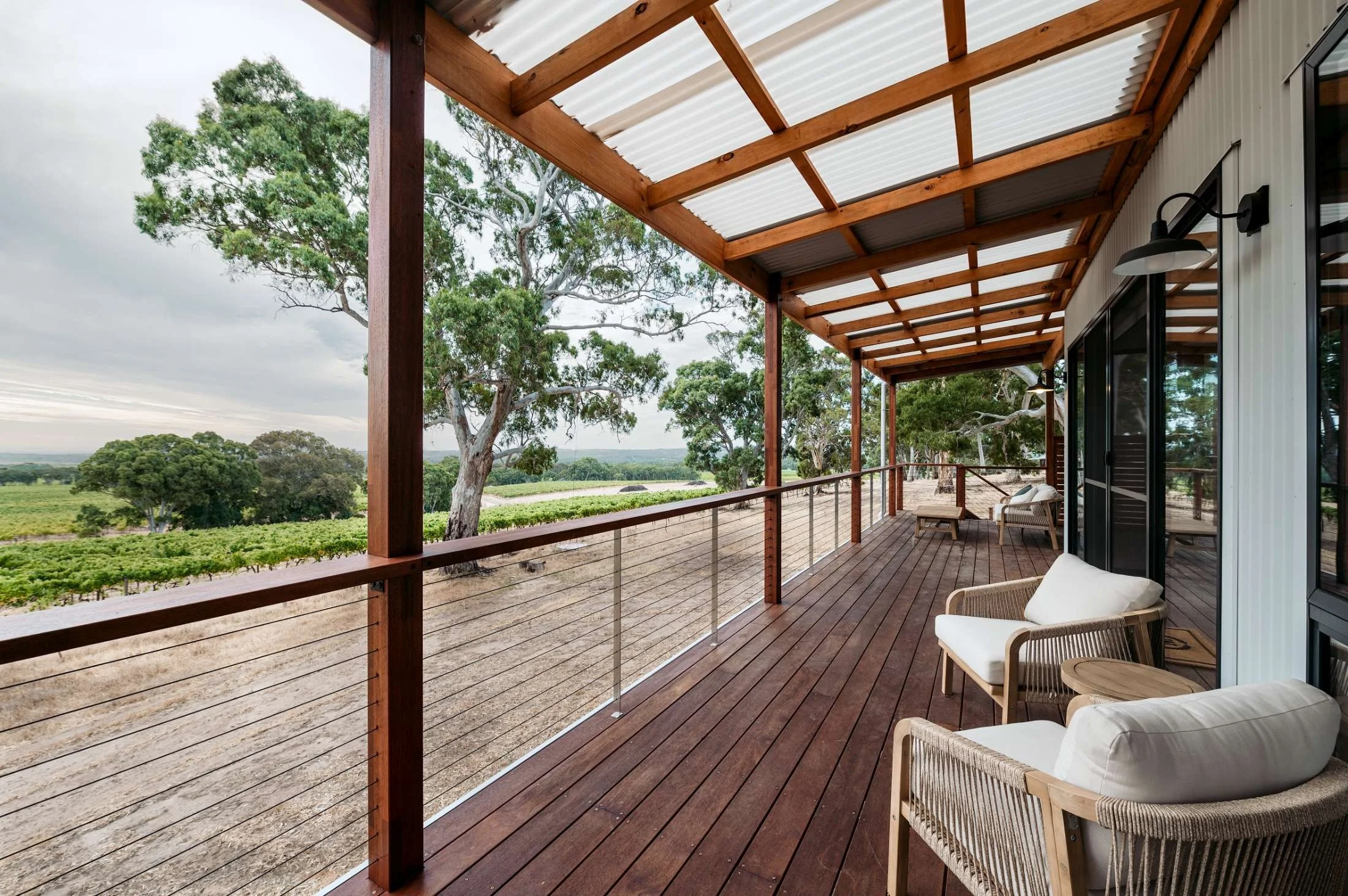 The image shows a spacious outdoor balcony with wooden flooring and a wooden and metal railing. There are cushioned chairs and a small table along the side of a building with large glass doors and black outdoor wall lights. The balcony overlooks green trees and farmland under a cloudy sky.