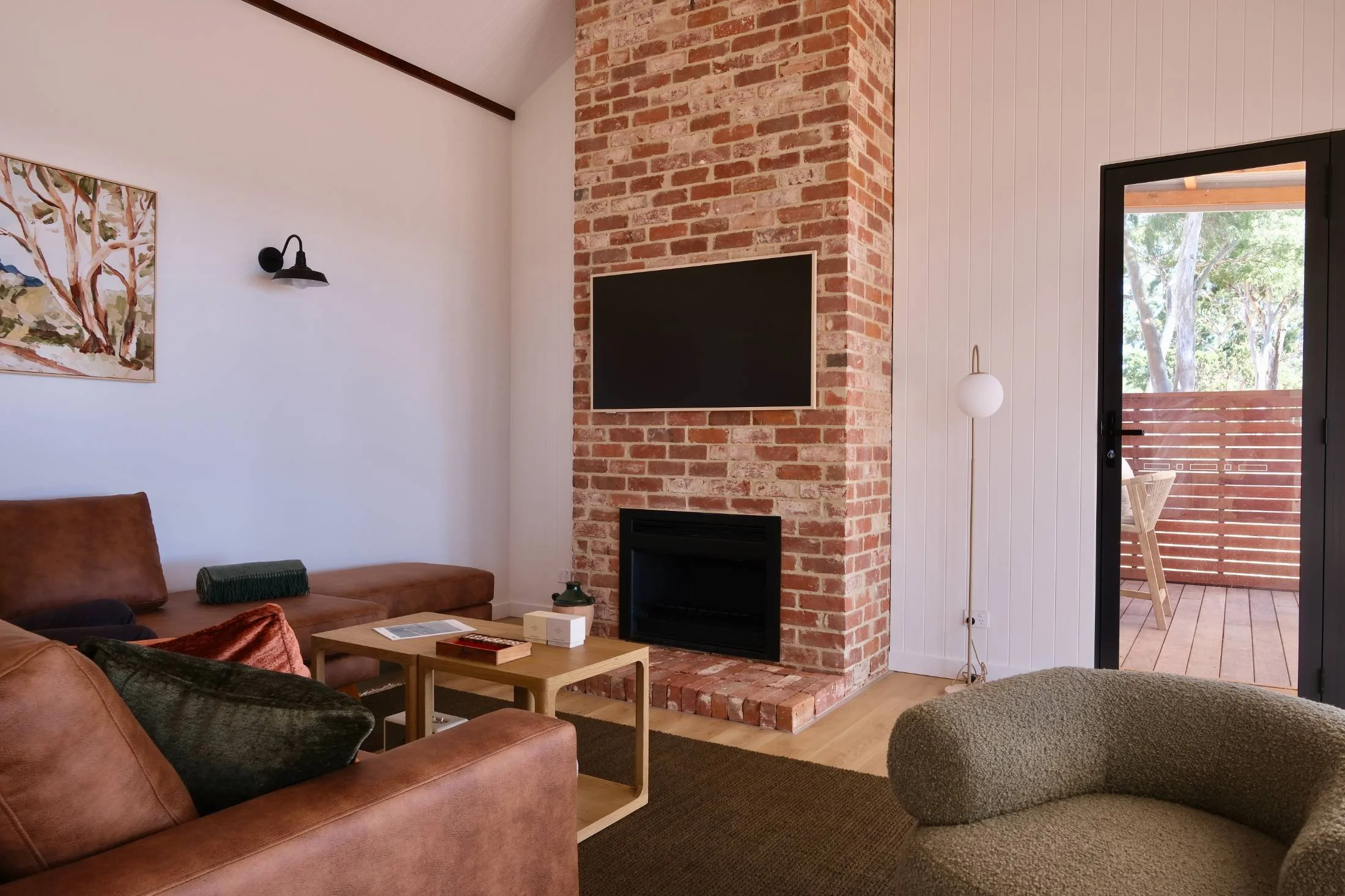 Living room with a brick fireplace, wall-mounted TV, brown leather sofa, and a sliding glass door leading to a wooden balcony with outdoor chairs.