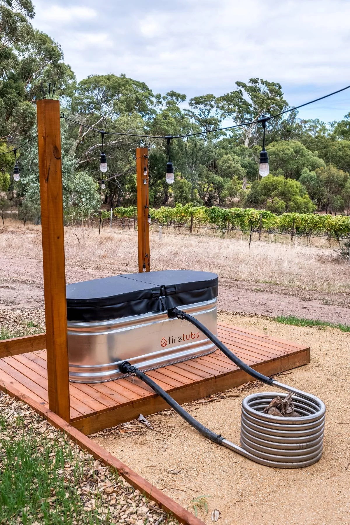 An outdoor hot tub with a black cover, connected to a copper coil heating system labeled 'fire tubs', on a wooden platform, with string lights overhead in a rural landscape with trees and vineyard in the background.