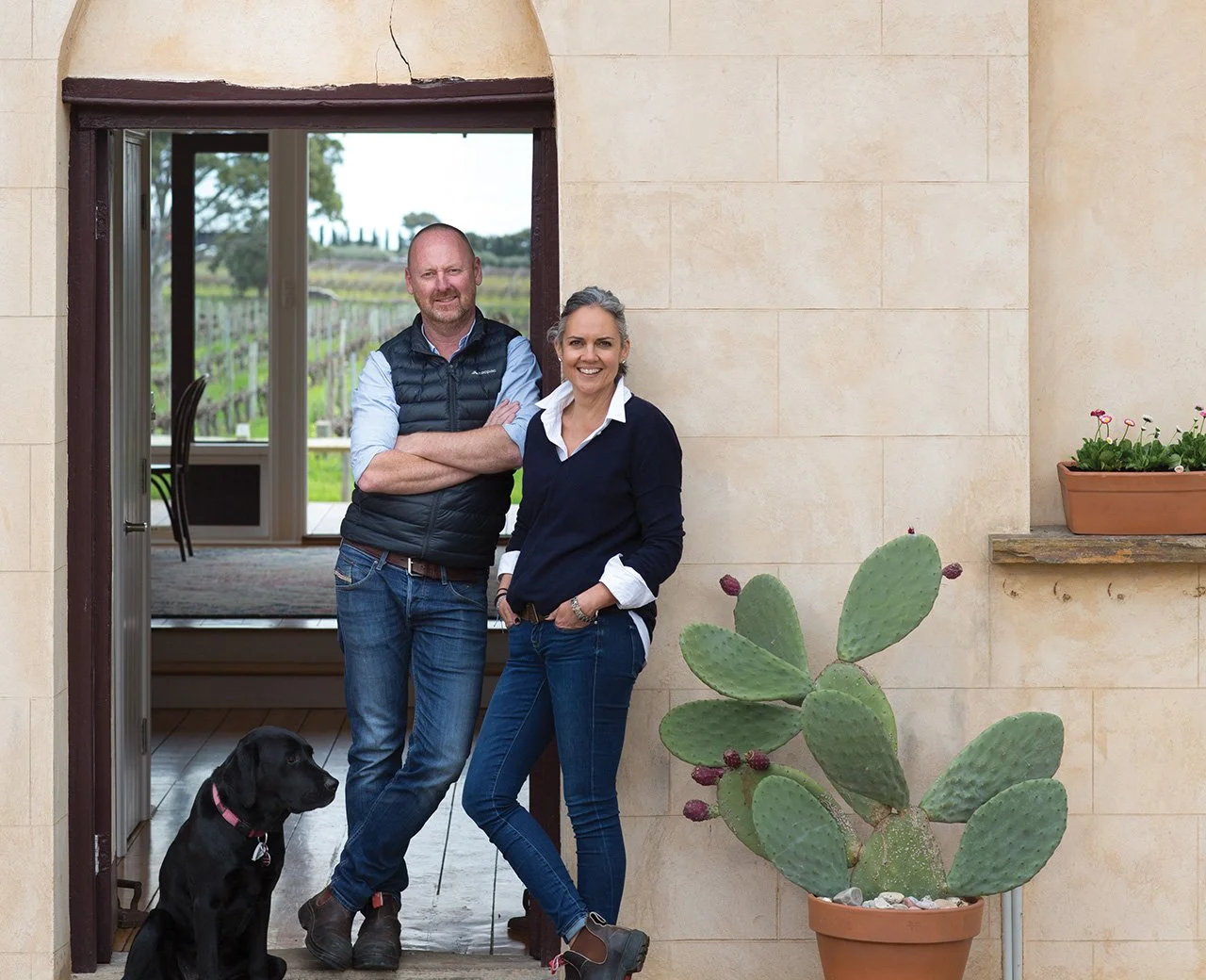 A man and woman standing in a doorway with a black dog and a cactus plant outside, with a vineyard visible through the open door.