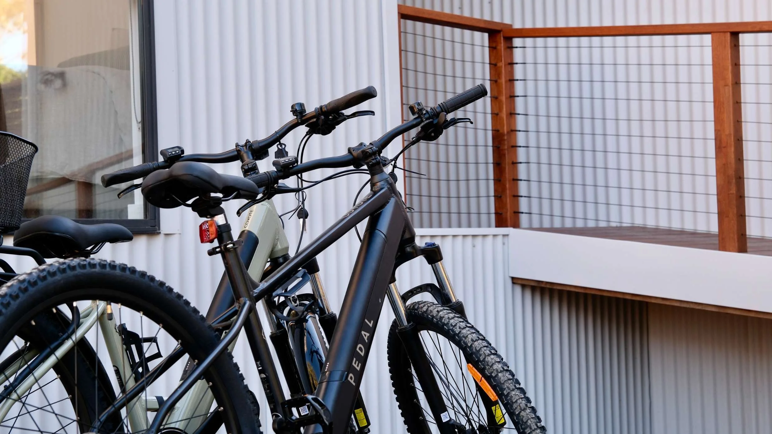 Two mountain bikes parked on a porch near a white wall with a window and a wooden railing.