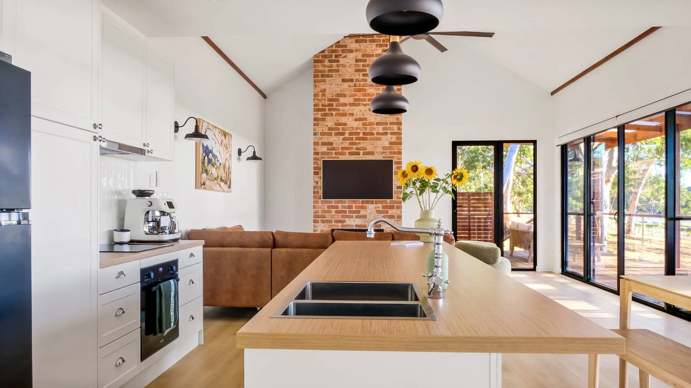 Open-concept kitchen and living room with white cabinetry, a brown leather sofa, brick accent wall with flat-screen TV, and sliding glass doors leading to an outdoor deck, with a vase of sunflowers on the kitchen island.