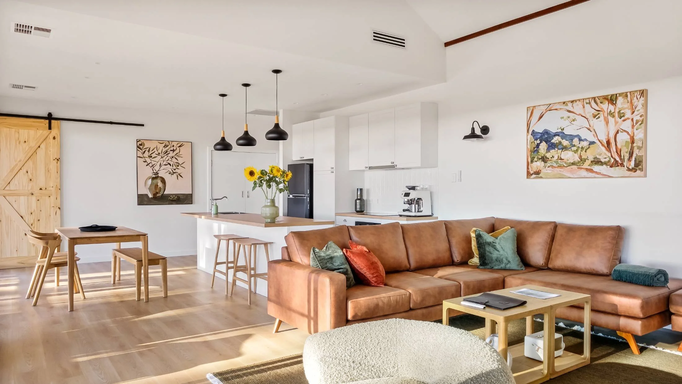 Living room with brown sectional sofa, colorful pillows, wooden coffee table, art on walls, dining area with wooden table and chairs, kitchen with white cabinets, black refrigerator, island with sunflower bouquet, black pendant lights.