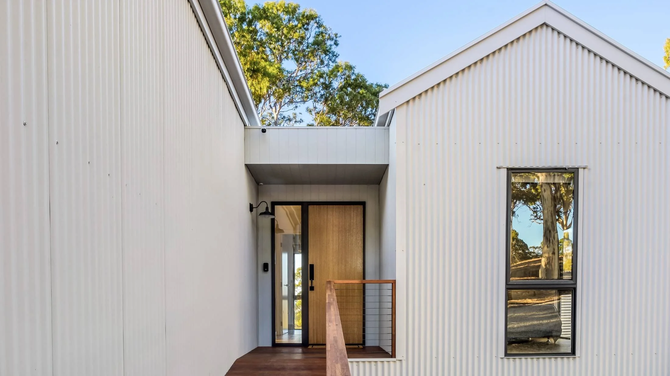 Modern house with white corrugated metal siding, wooden door, and large window reflecting trees, with a small front porch and railing, set against a background of greenery and blue sky.