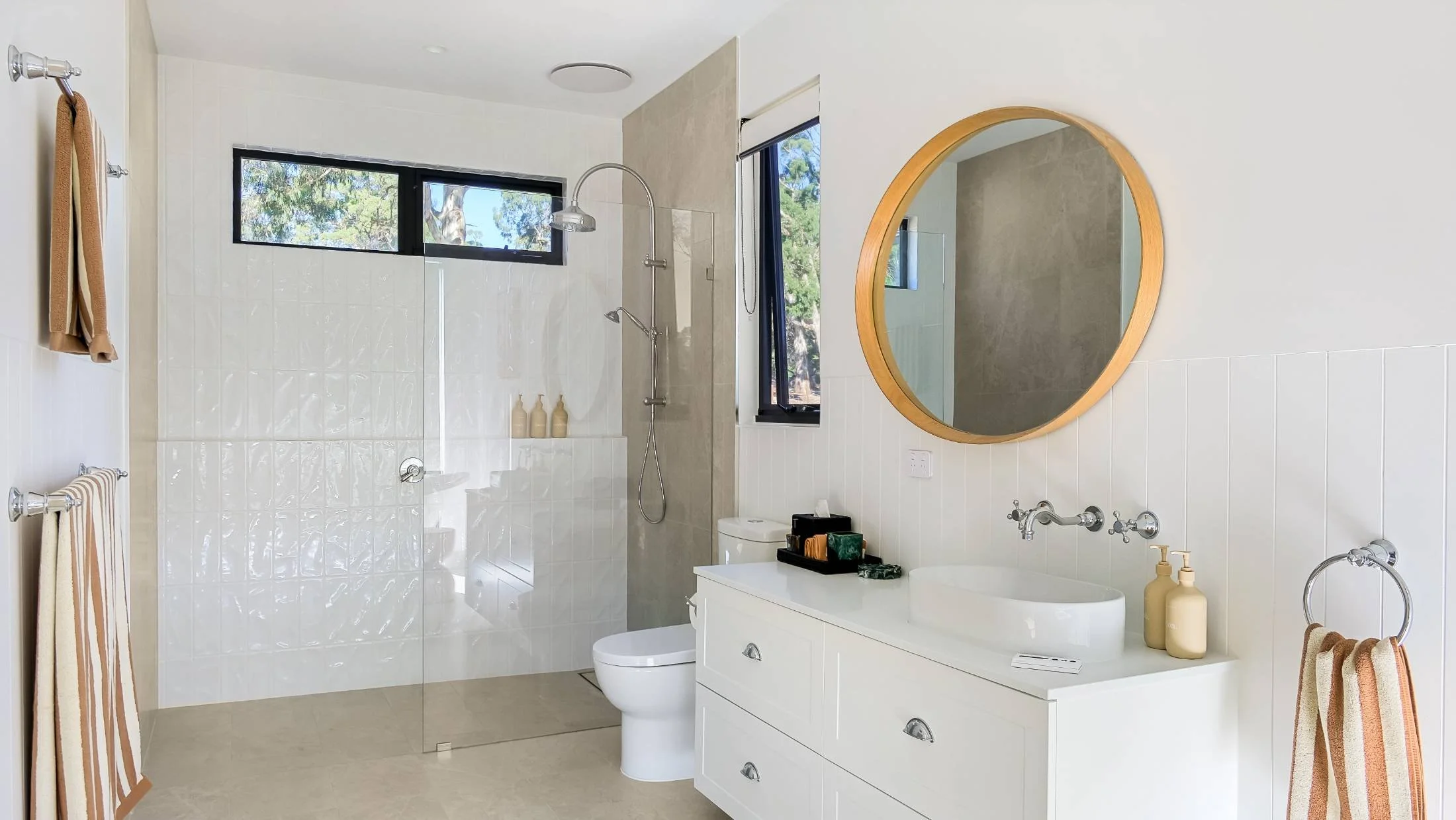 Modern bathroom with white walls, a vanity with a round mirror, a vessel sink, bottles, a toilet, and a glass-enclosed shower with a window and beige tiles.