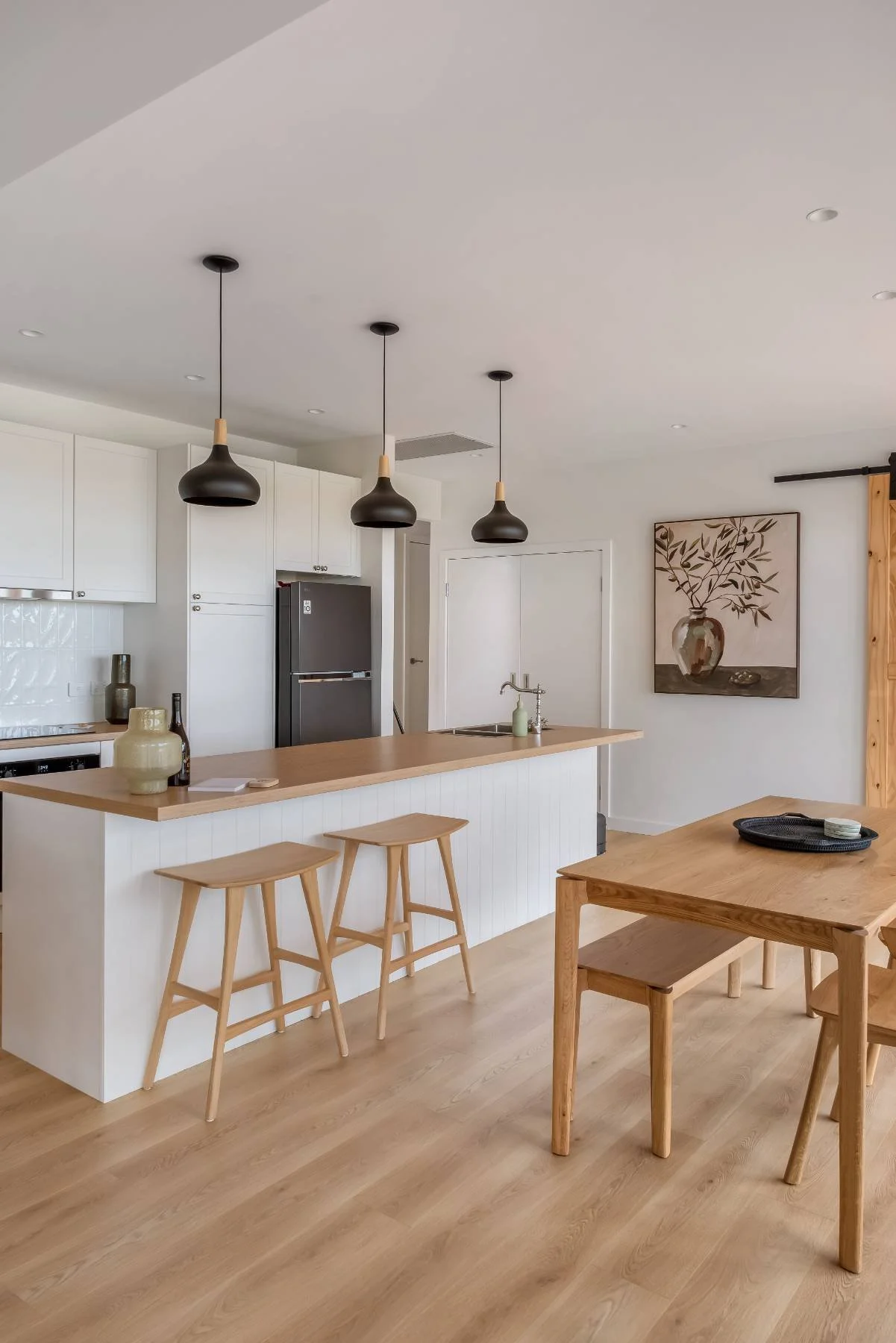 Modern kitchen and dining area with white cabinets, black refrigerator, wooden kitchen counter with two stools, wooden dining table with chairs, decorative vases, and a framed botanical wall art.