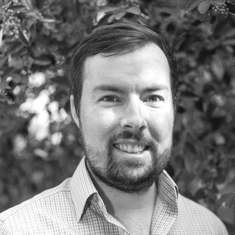 Black and white photo of Brett Findlaey, smiling, wearing a collared shirt, with a blurred leafy background.