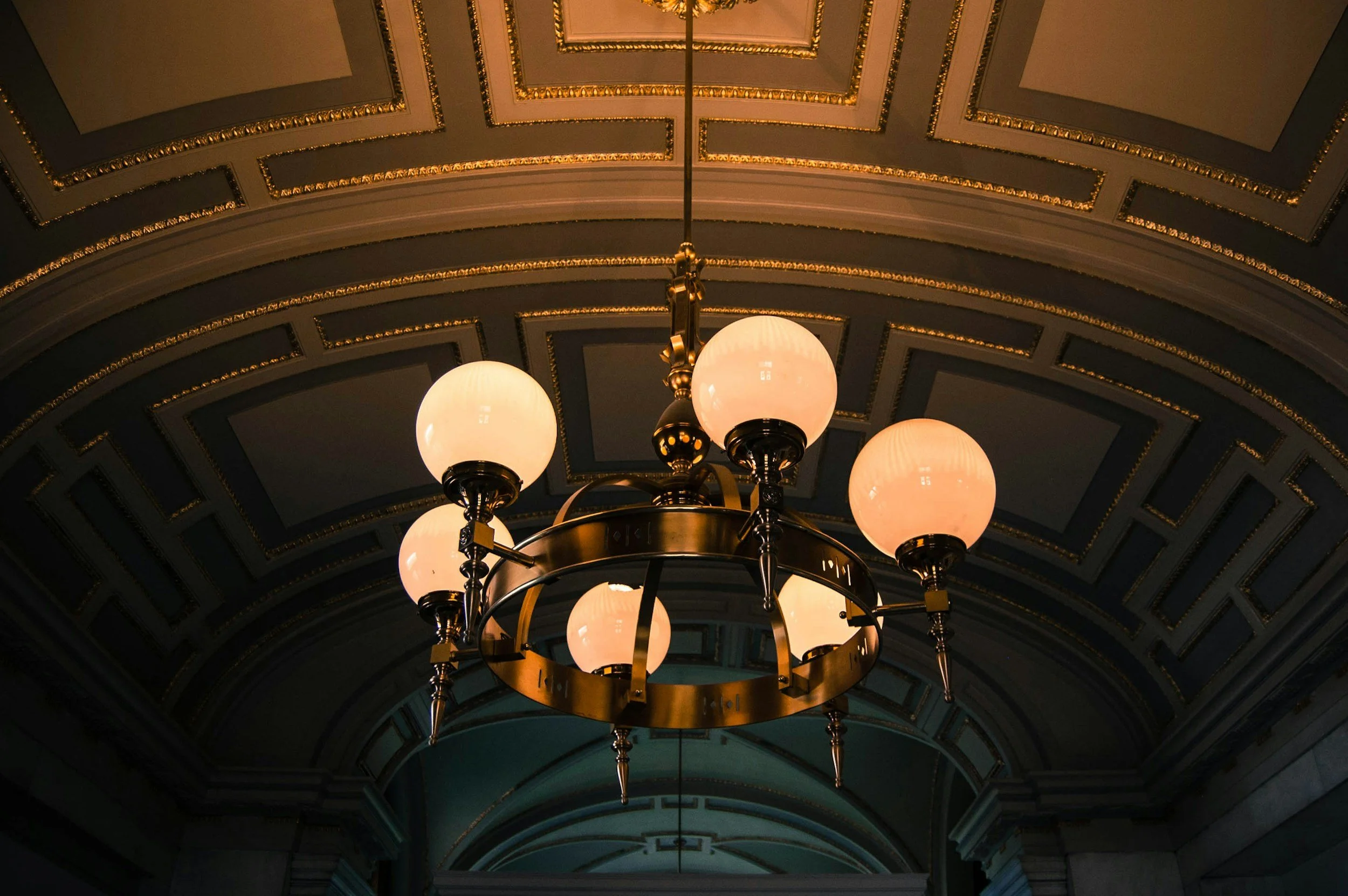 A chandelier with six spherical white lights hangs from a decorative ceiling with gold accents and geometric patterns.