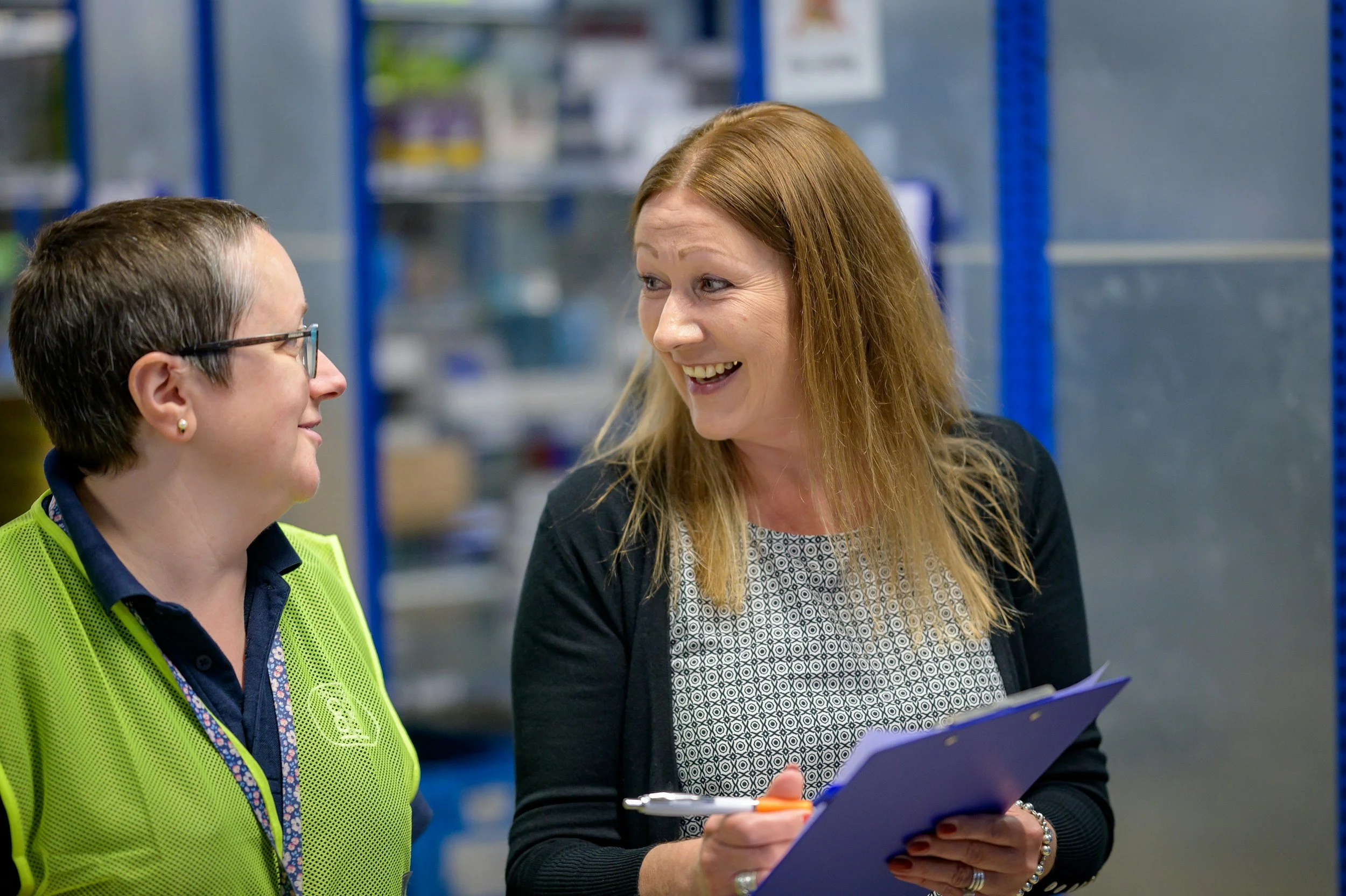 Two women having a conversation in a warehouse, one holding a clipboard and pen, both smiling.