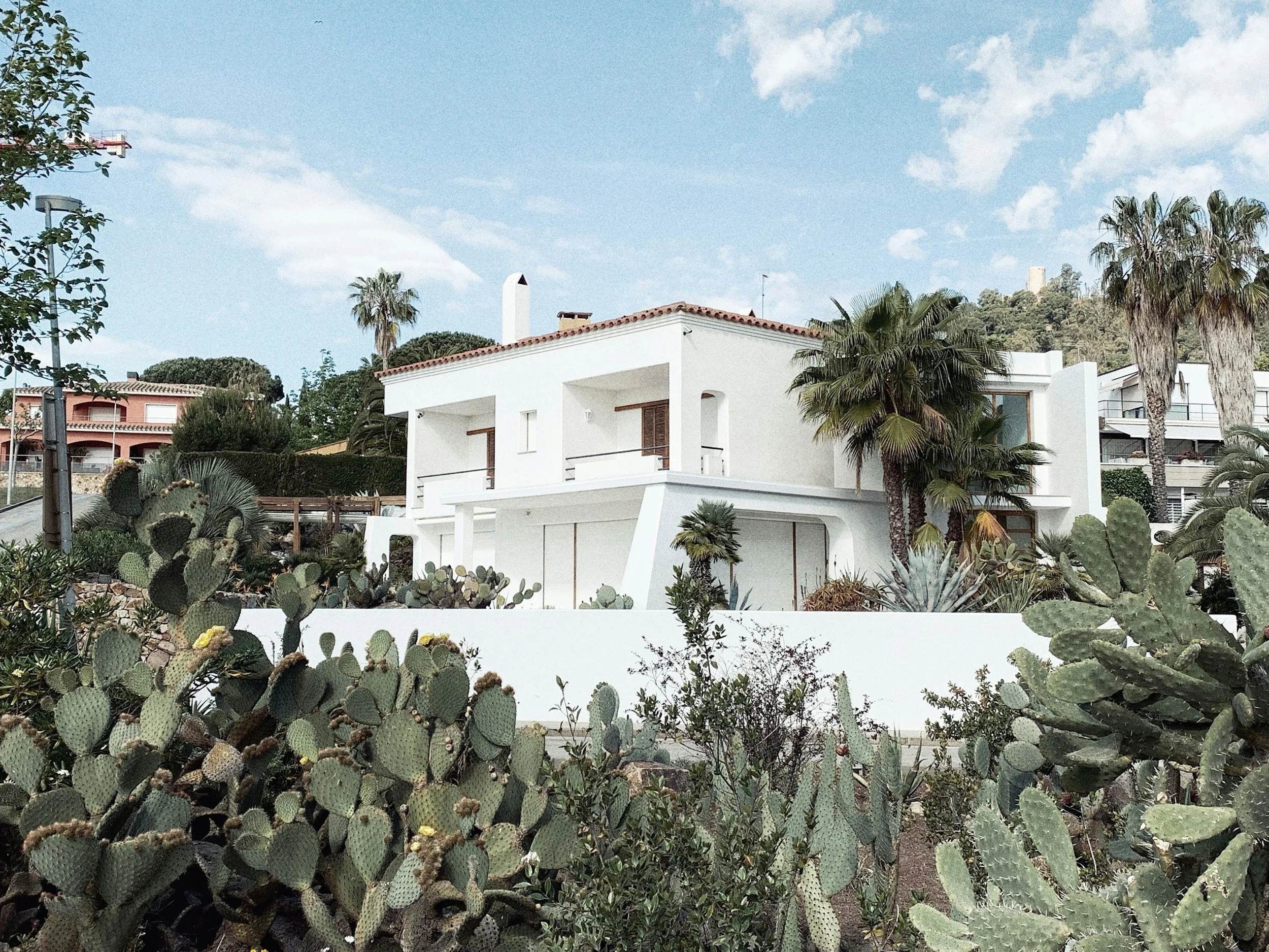 A modern white house with a flat roof, surrounded by a desert landscape with cacti and palm trees, under a partly cloudy sky.