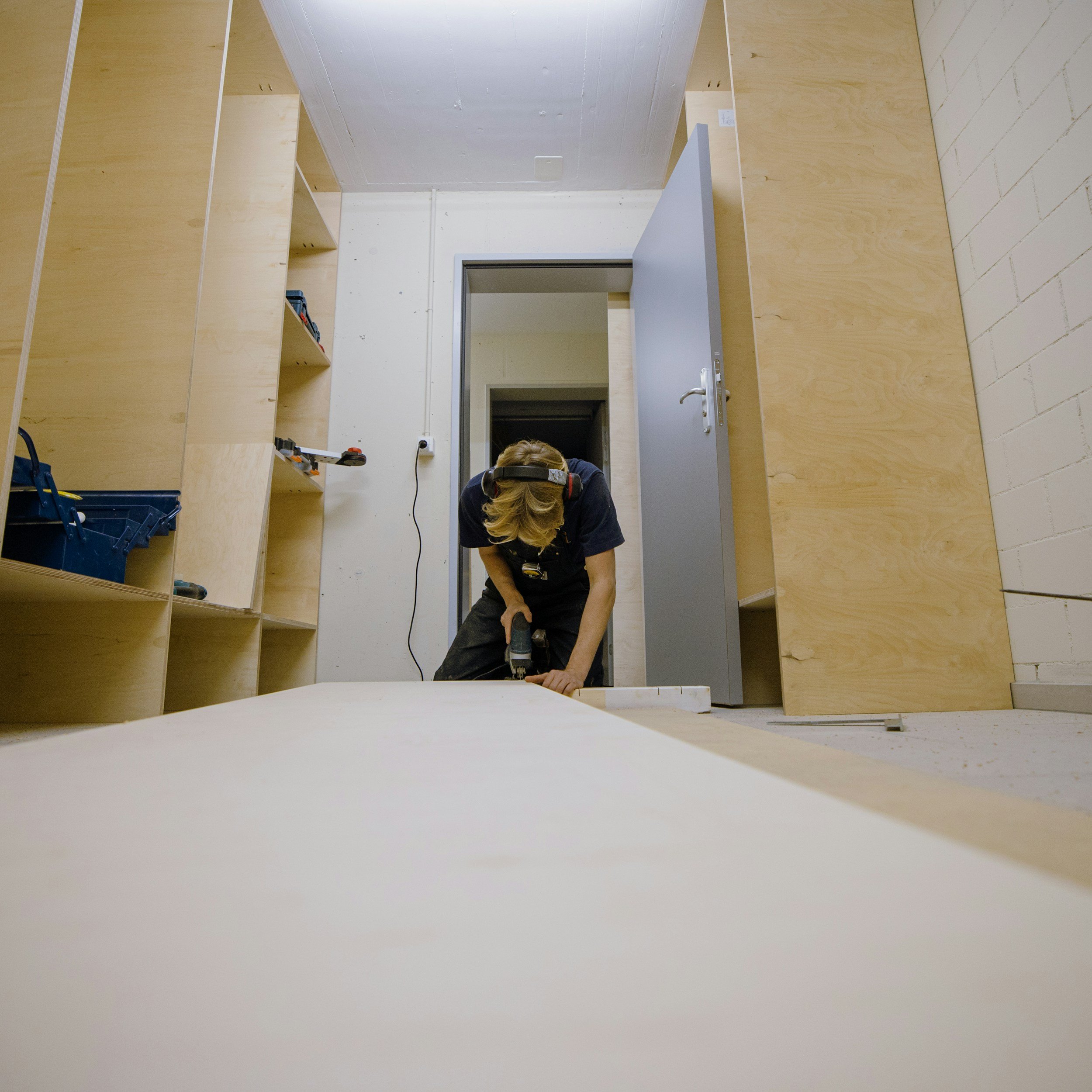 A person working on woodworking in a workshop, leaning over a large piece of wood, wearing safety headphones, with shelves and tools around.