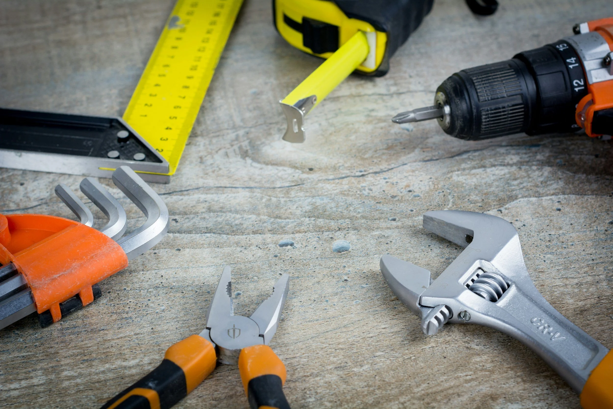 Tools on a wooden surface, including a tape measure, screw gun, pliers, adjustable wrench, and a cordless drill.
