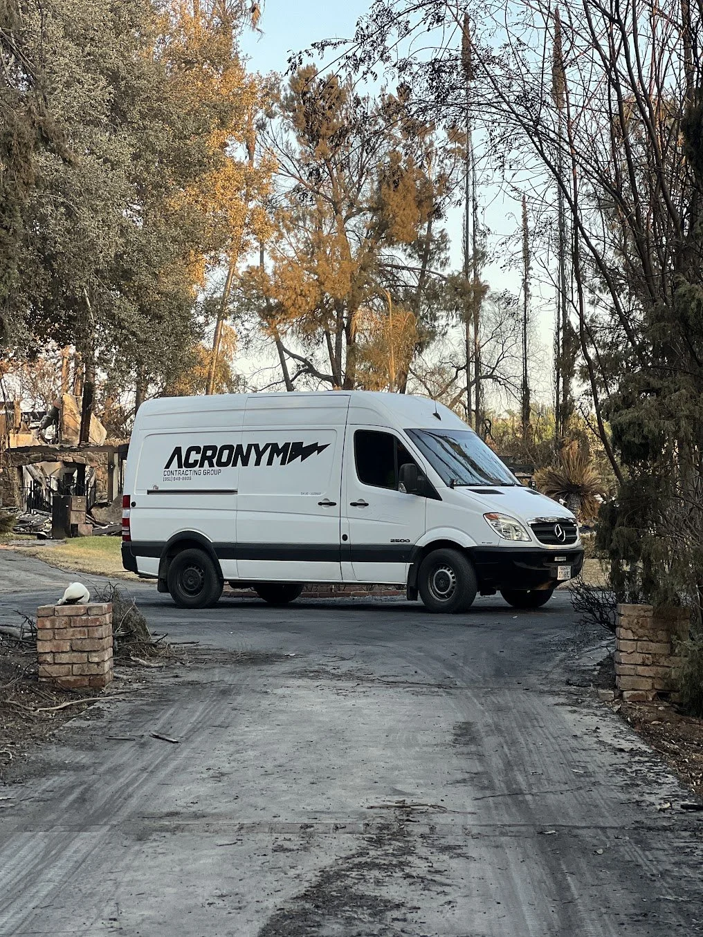 A white Mercedes-Benz sprinter van with contractor branding, parked on a dirt driveway surrounded by trees and damaged structures.