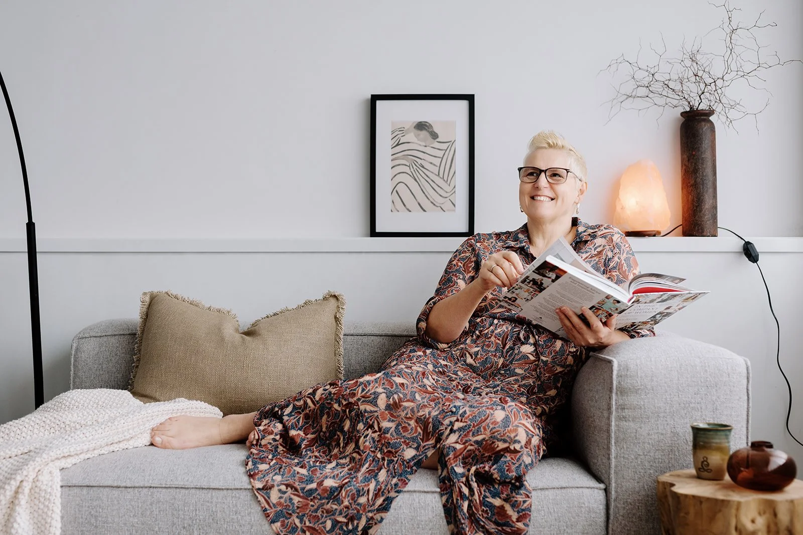 Owner, Marie, smiling woman with short blonde hair and glasses sitting on a gray sofa, reading a magazine in a cozy, modern living room.