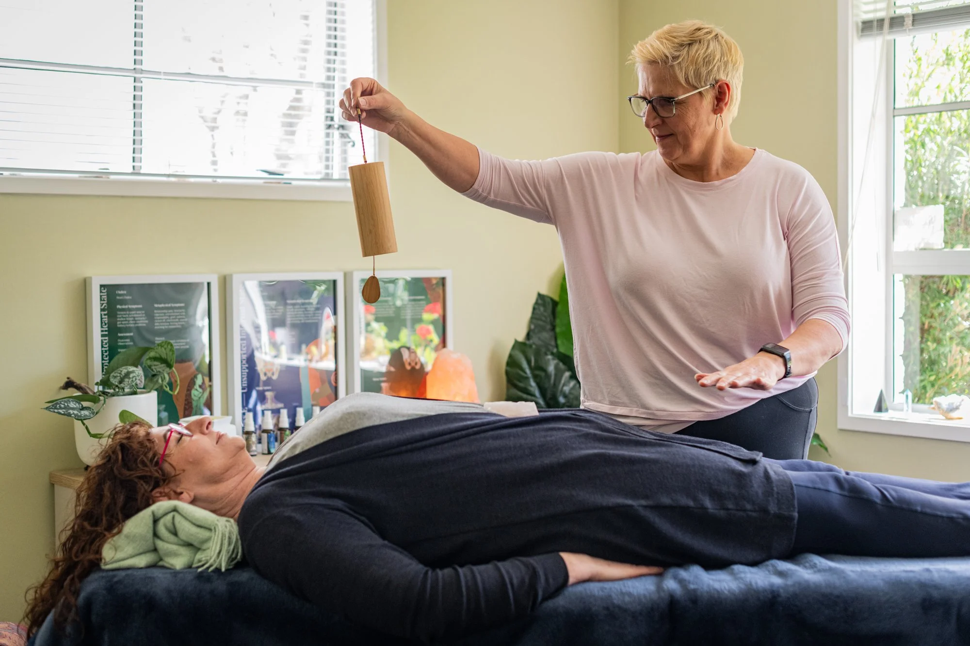 A woman lying on a massage table receiving energy healing from a practitioner holding a wooden pendulum over her abdomen in a bright room with green walls and large windows.