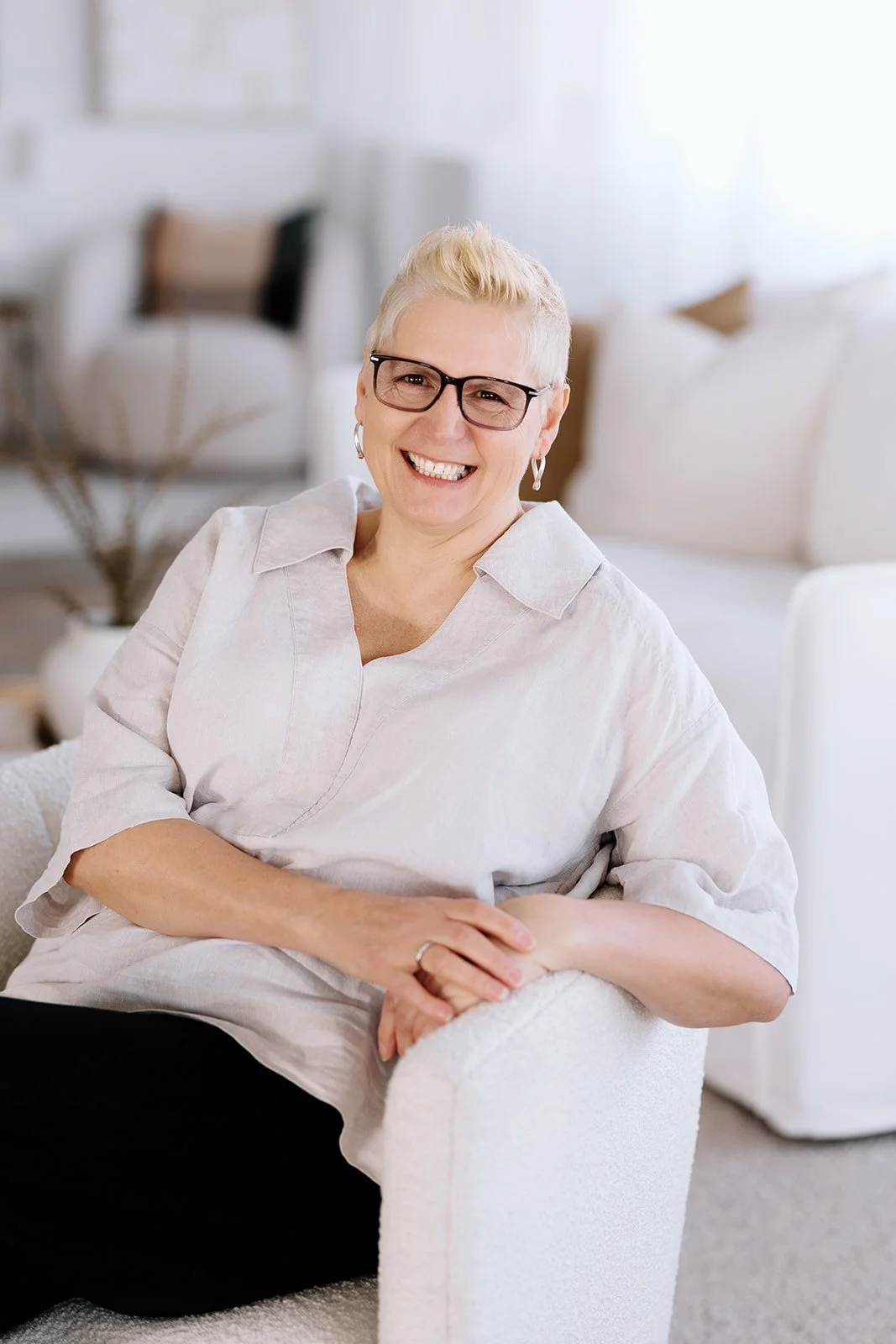 Owner, Marie smilingwith short blonde hair, wearing glasses and a light-colored blouse, sitting on a white couch in a bright, modern living room.
