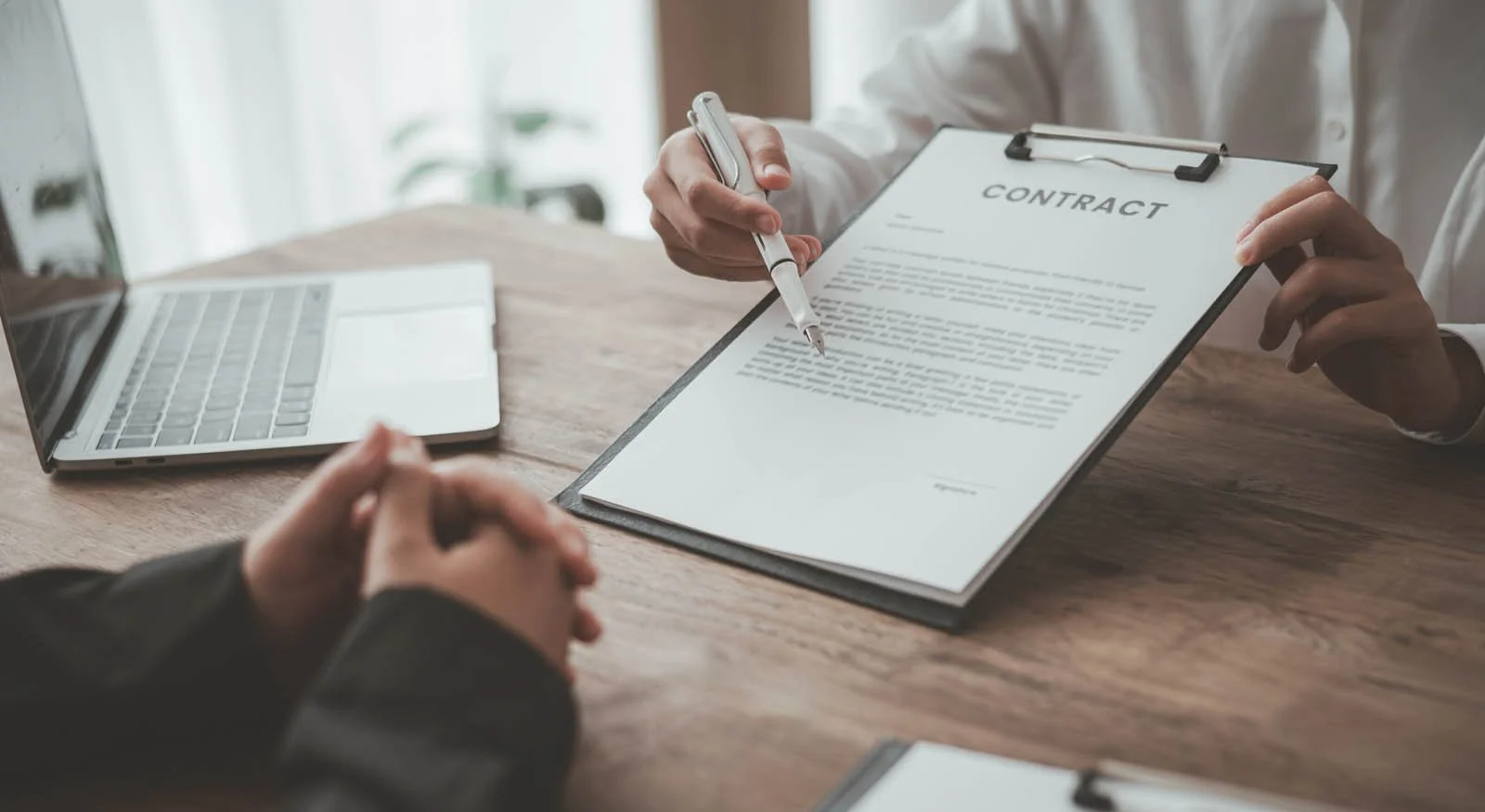 Kuvassa henkilö allekirjoittaa sopimusta, vieressä on toinen henkilö ja avattu kannettava tietokone.