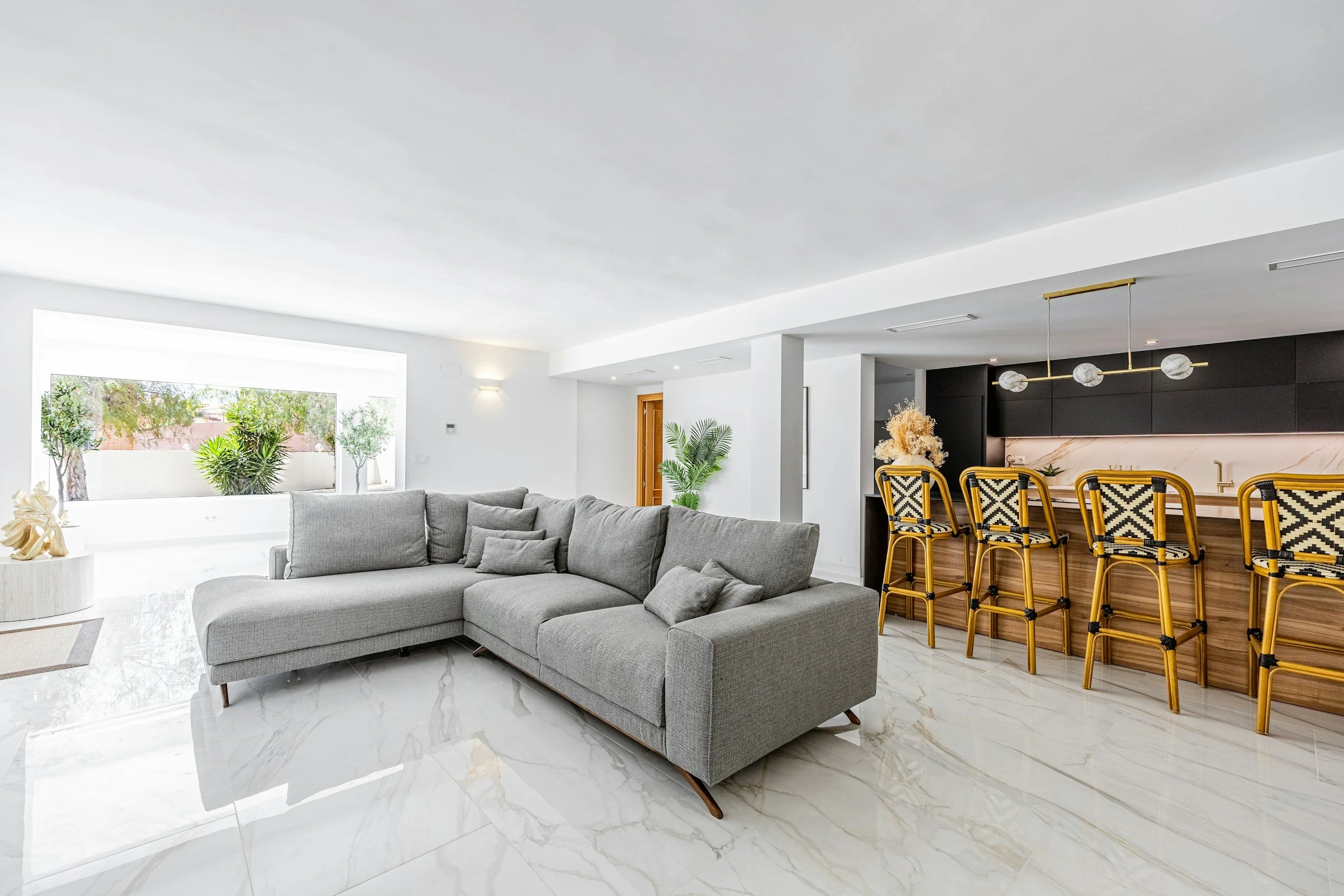 Modern living room with a large gray sectional sofa, marble floors, a window with greenery outside, and a kitchen bar area with black cabinets, marble backsplash, and yellow and black patterned bar chairs.